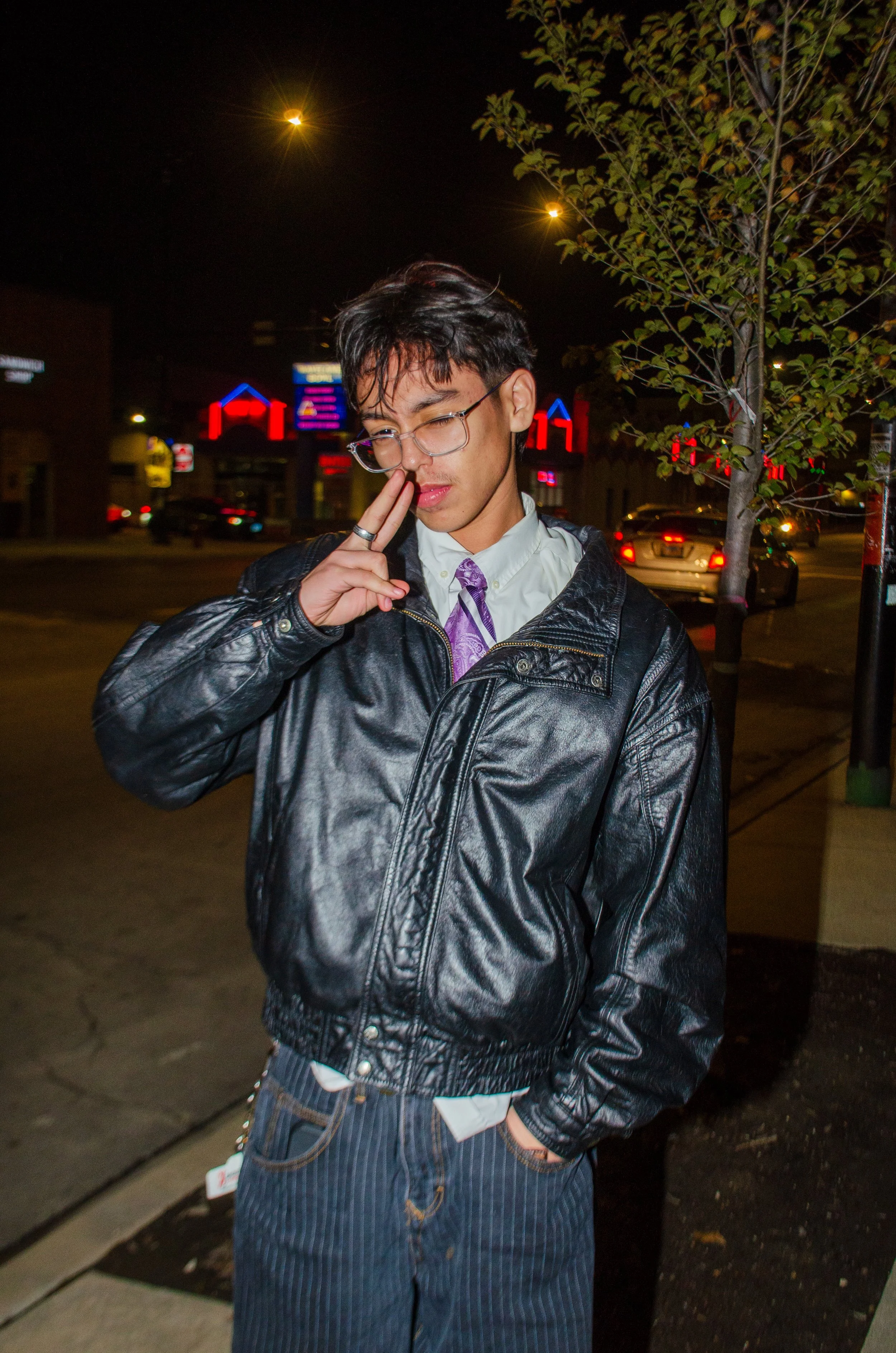A young man with glasses, black hair, and light skin, standing on a city street at night. He is wearing a black leather jacket, a white shirt, a purple tie, and pinstriped pants. He is touching his nose with his right hand and has a thoughtful or pla