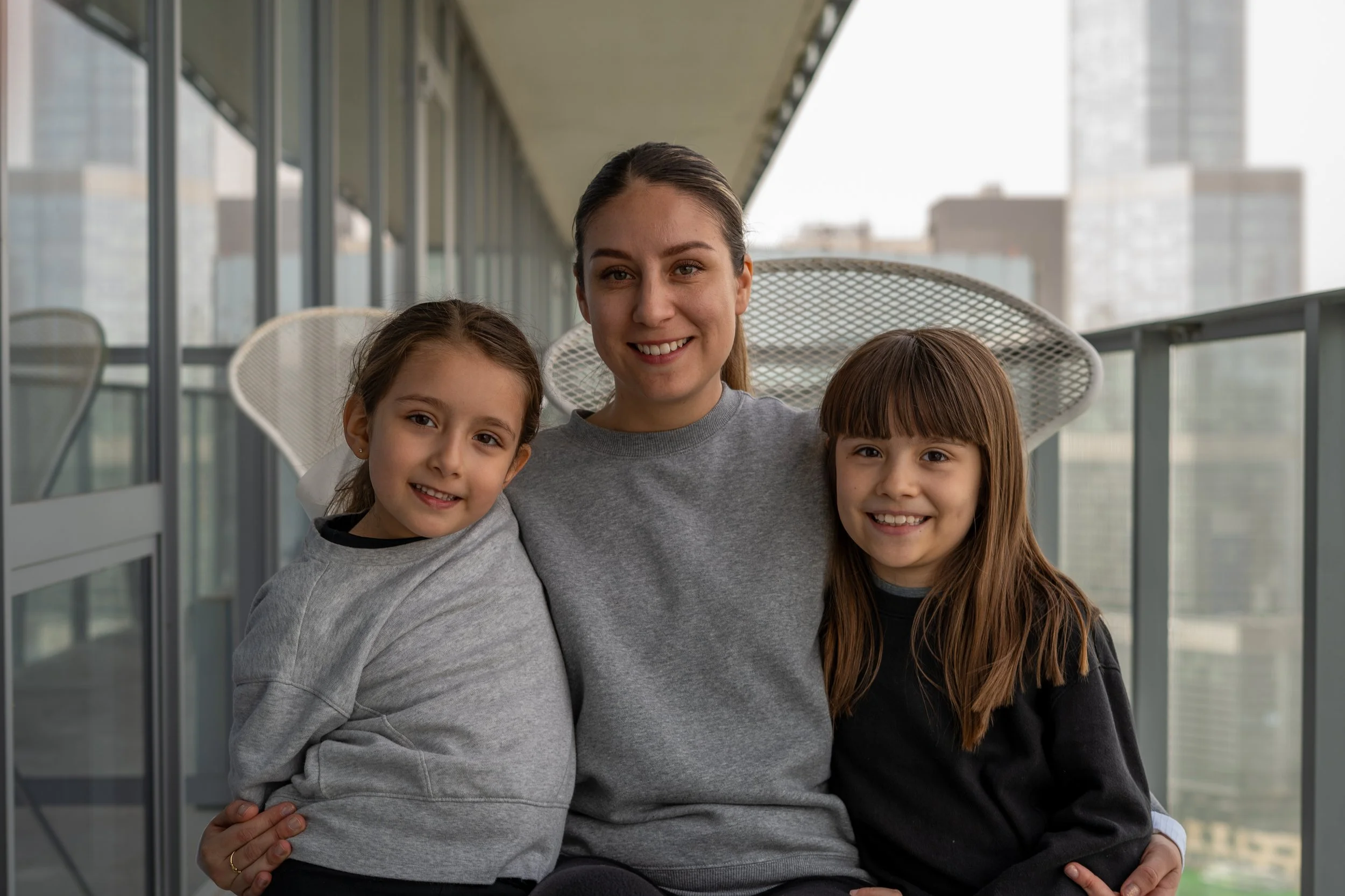 A woman with brown hair in a ponytail smiling and sitting on a balcony with two young girls, one with dark brown hair and the other with light brown hair, all wearing casual clothing and smiling at the camera.