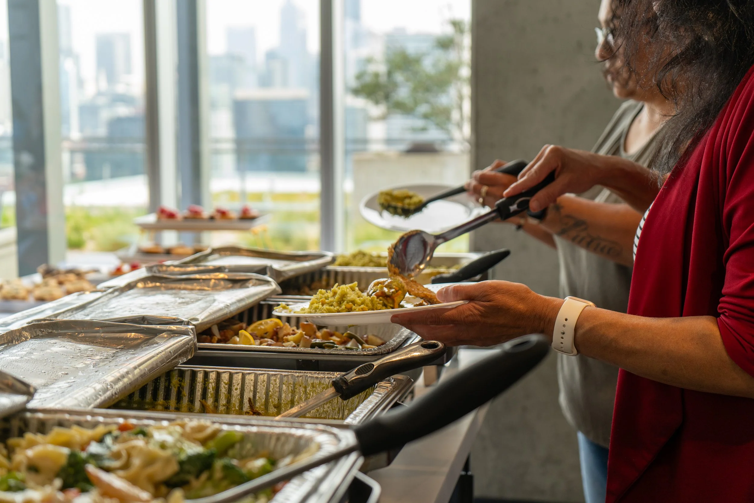 People serving themselves food from a buffet table inside a building with large windows, showing a cityscape in the background.