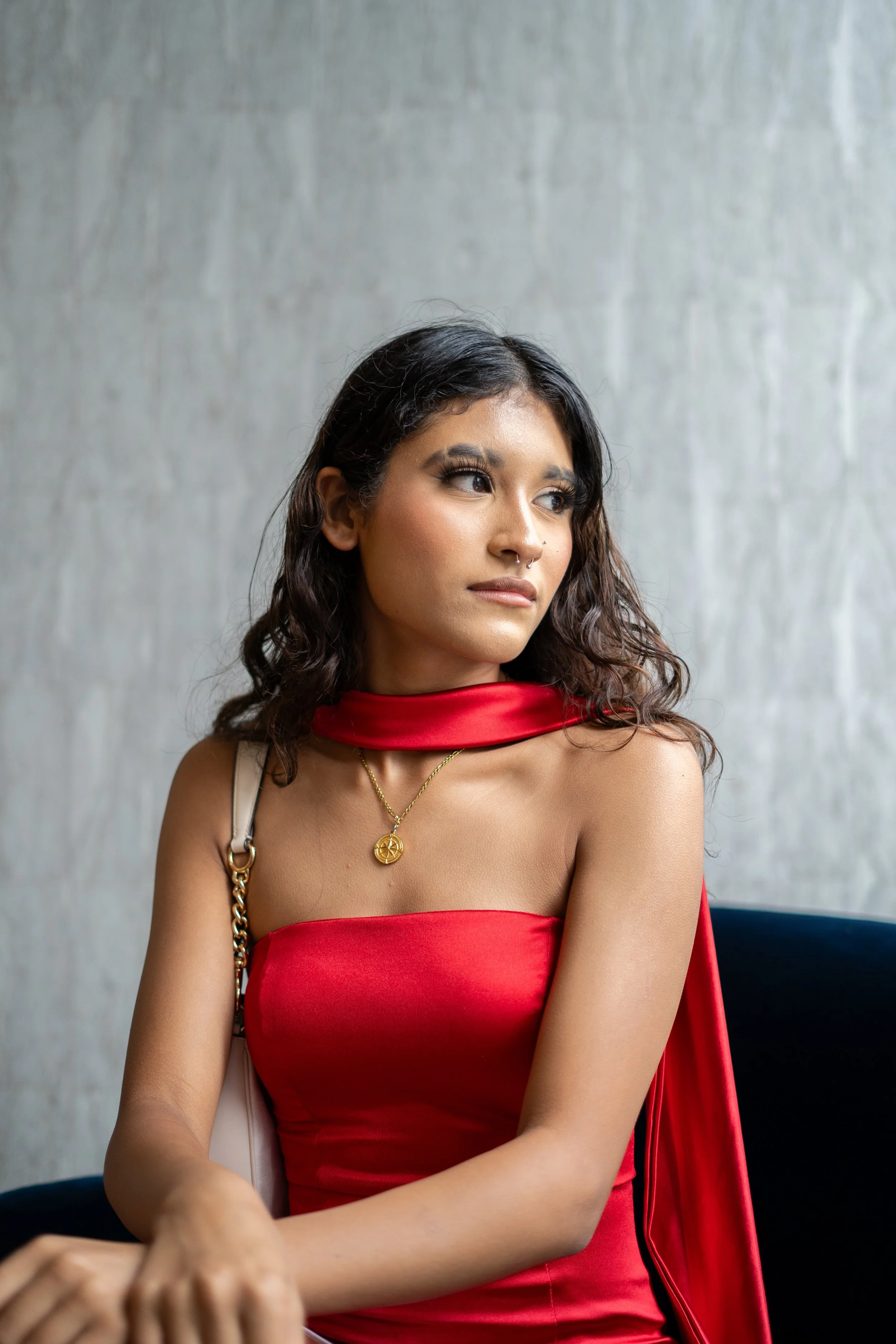 Young woman with long dark curly hair, wearing a red strapless dress, a gold necklace with a circular pendant, and a septum piercing, sitting against a plain gray wall, looking to her left.