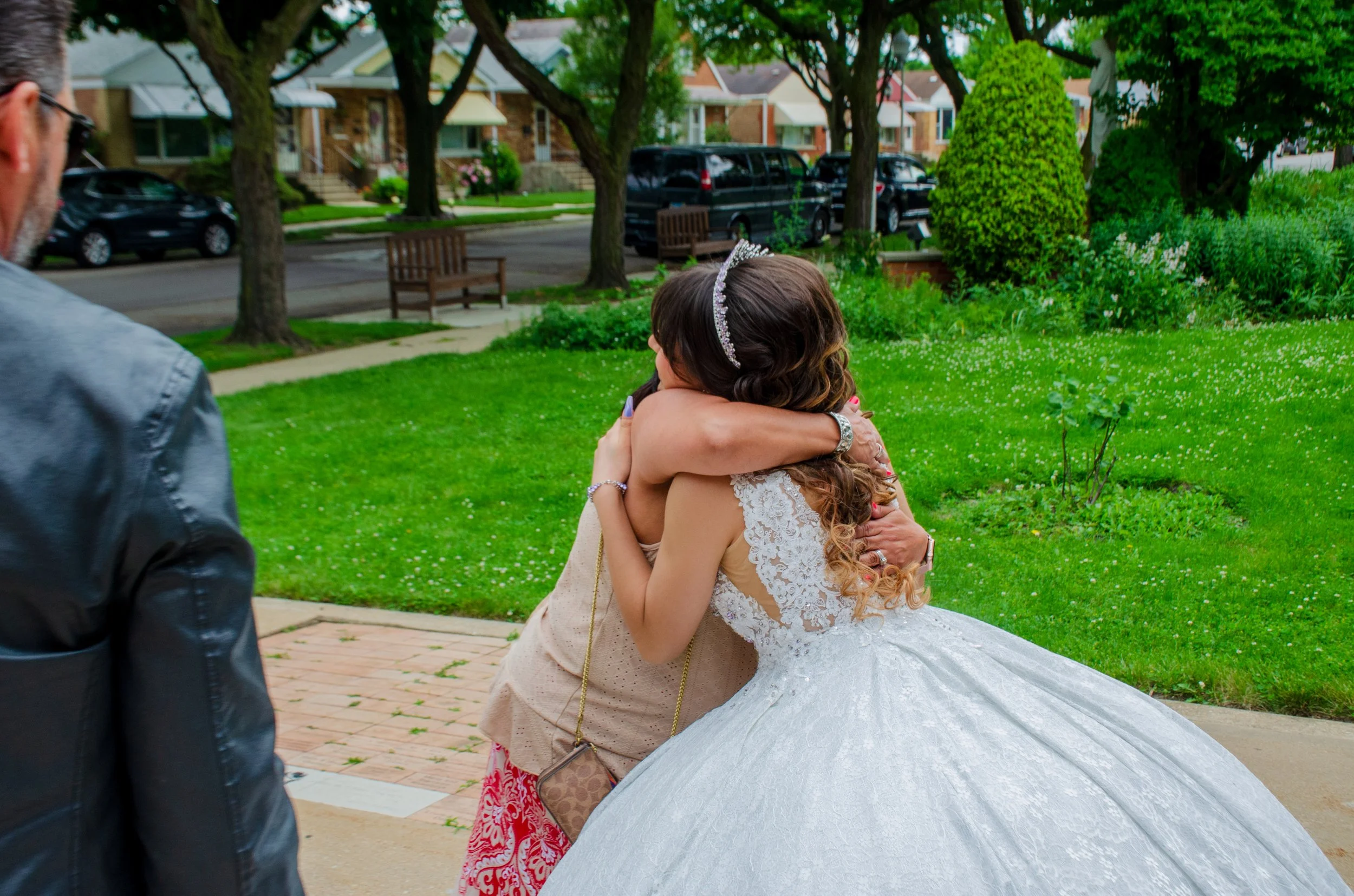 A bride in a white wedding gown wearing a tiara is hugging an older woman outside in a park, with a large umbrella on the ground nearby. A man in a gray jacket is partially visible on the left.