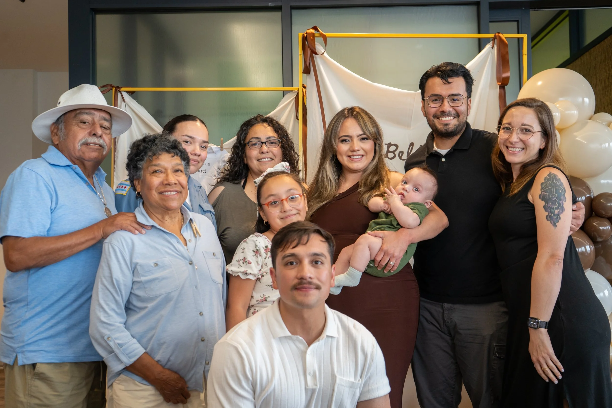 A group of diverse people smiling and posing for a photo at an indoor event. There are balloons and decorative drapes in the background.