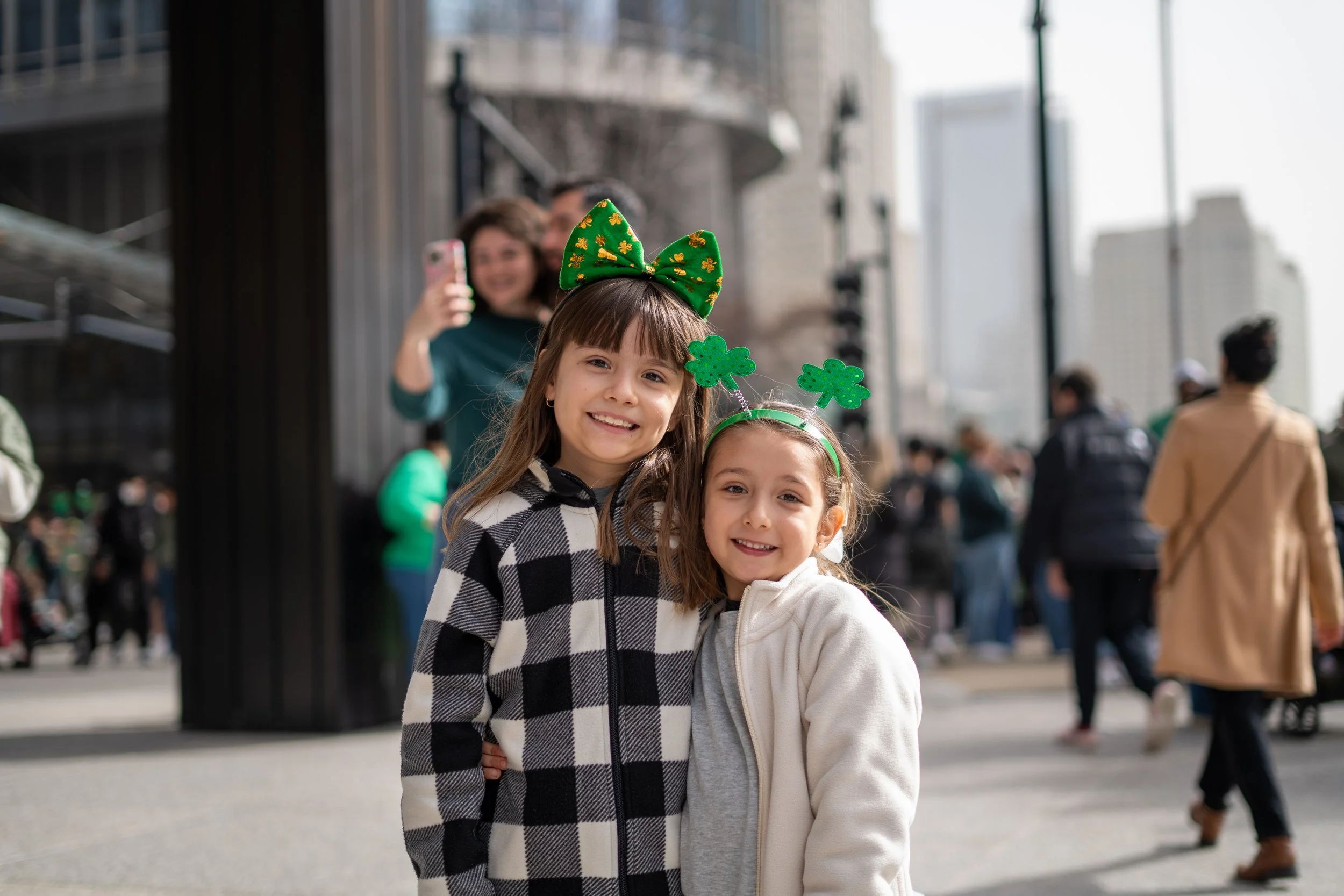 Two young girls dressed in festive attire with green shamrock headbands smiling in a city street, with a woman taking their picture in the background.
