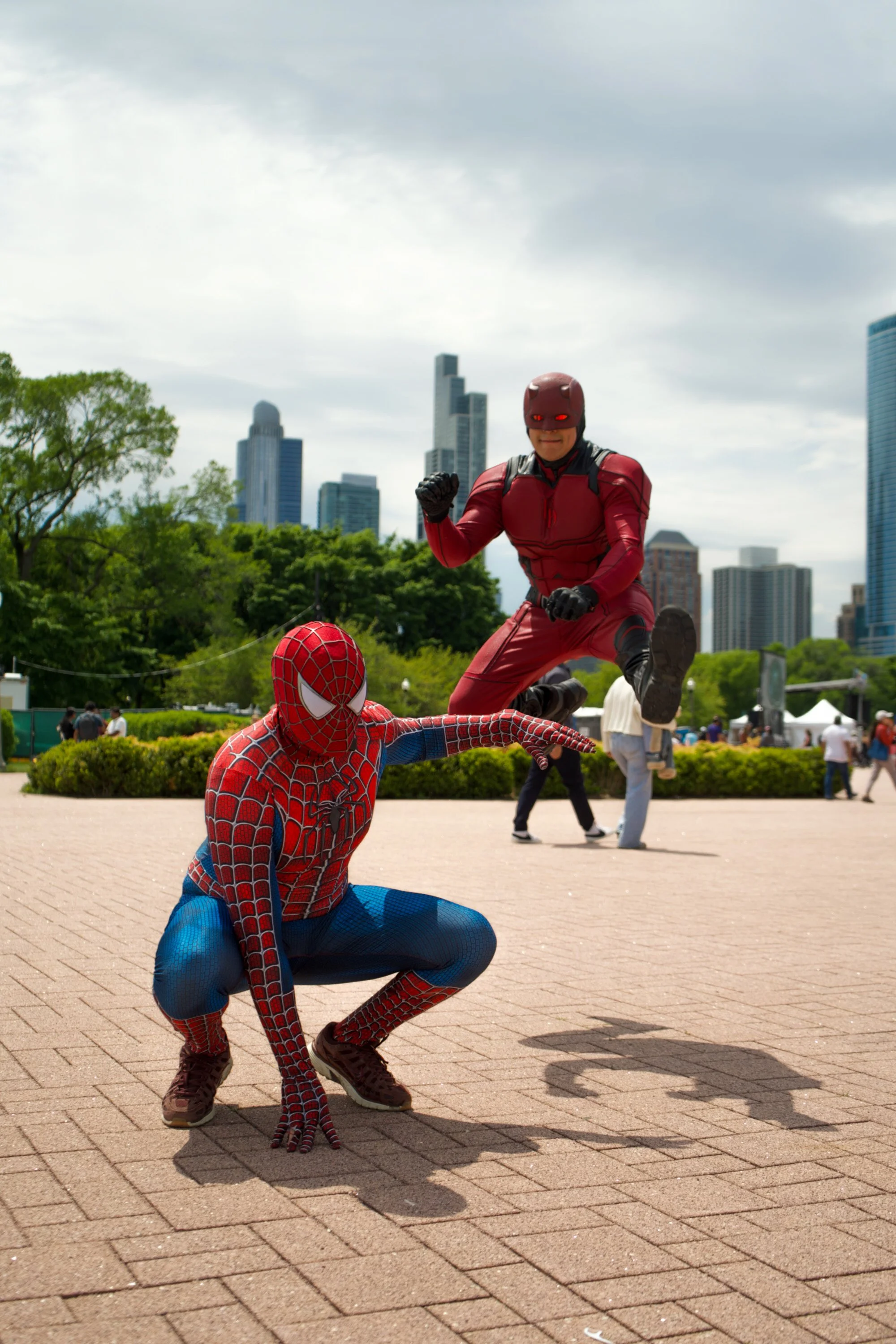 Two people dressed as Spider-Man and Daredevil in superhero costumes, posing outdoors in a park with a city skyline in the background.