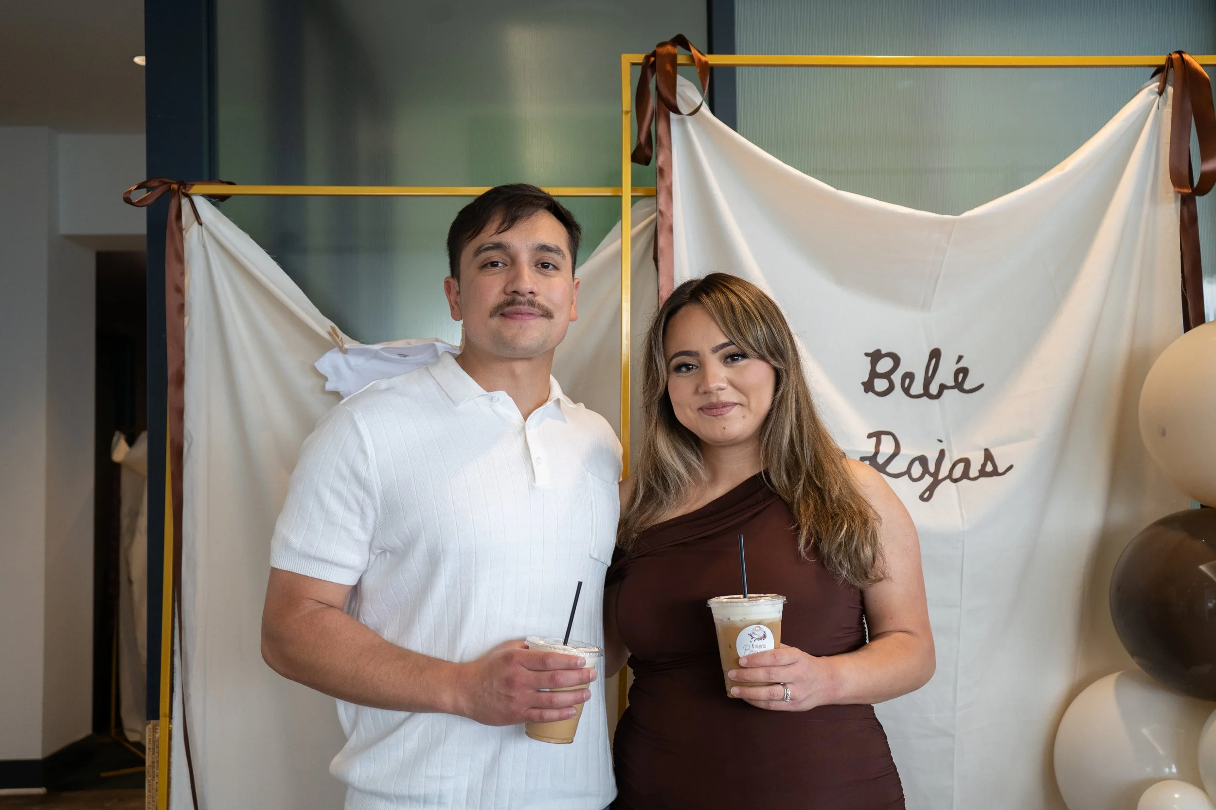 A man and a woman standing together, holding iced coffee drinks, in front of a decorative backdrop with balloons and a white cloth with dark writing.