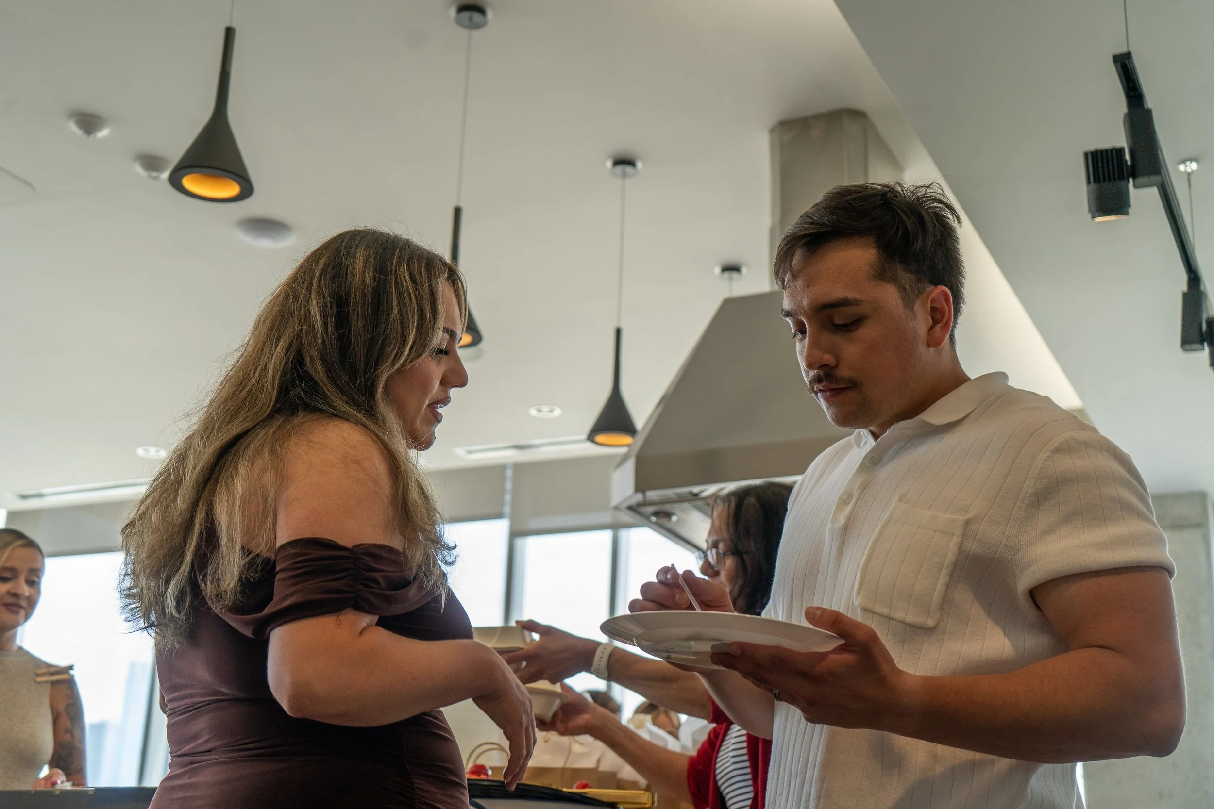 A man and woman are standing close together indoors, sharing an intimate moment as the man presents her with a necklace on a chain. The woman has long, wavy blonde hair and is wearing a dark brown dress, while the man has short dark hair and is weari
