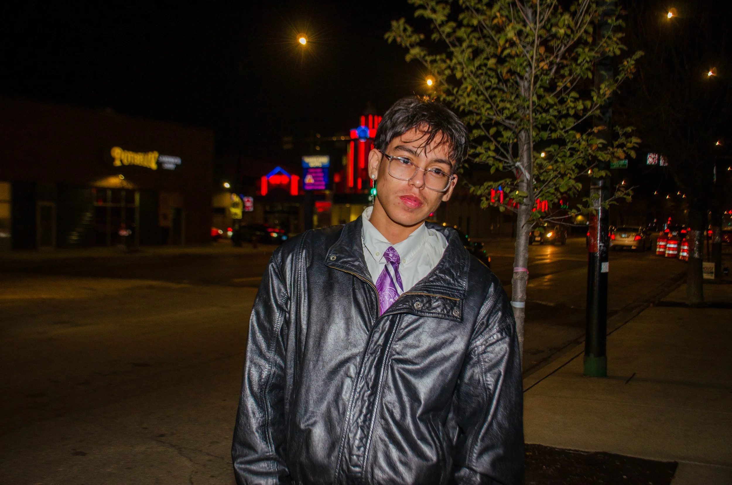 Young person with short black hair, glasses, and earrings standing on a city street at night, wearing a leather jacket, white shirt with purple tie, with illuminated signs and traffic in the background.