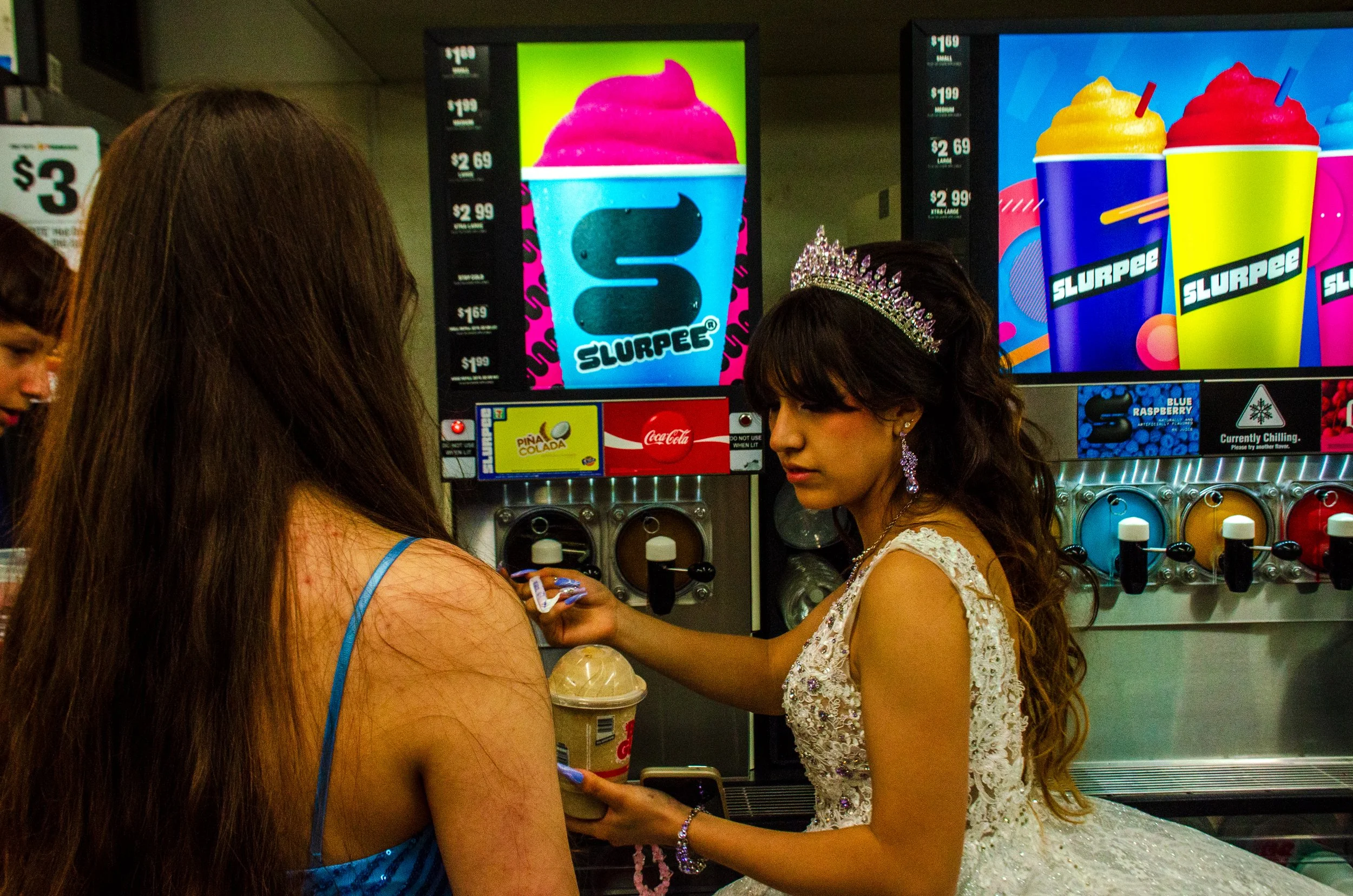 A young girl wearing a tiara and earrings is buying a pink cotton candy from a machine at a fair or amusement park, with other people visible around.
