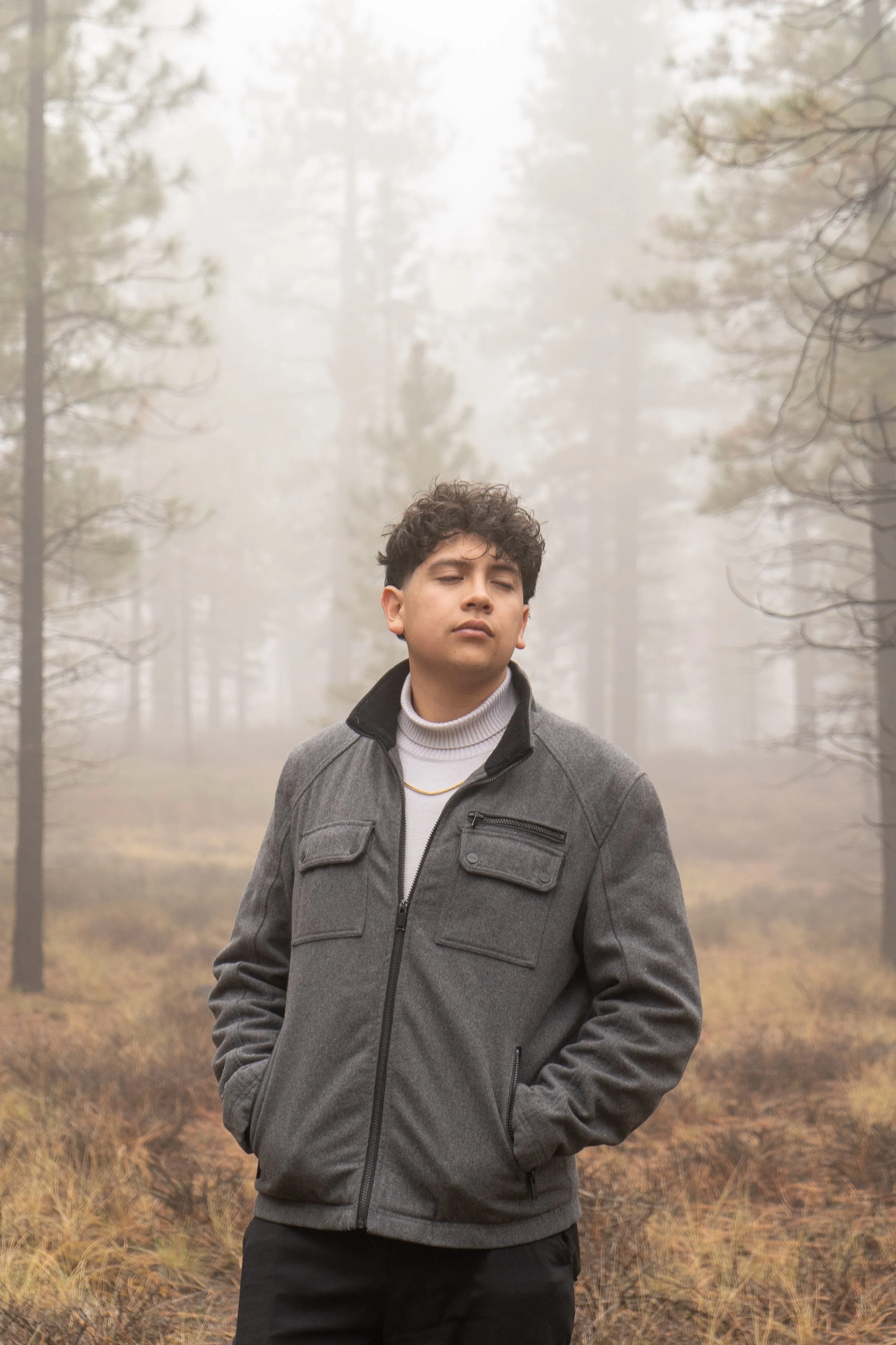 A young man with curly hair stands outdoors in a foggy forest, wearing a gray jacket with a quarter zip and black pants.