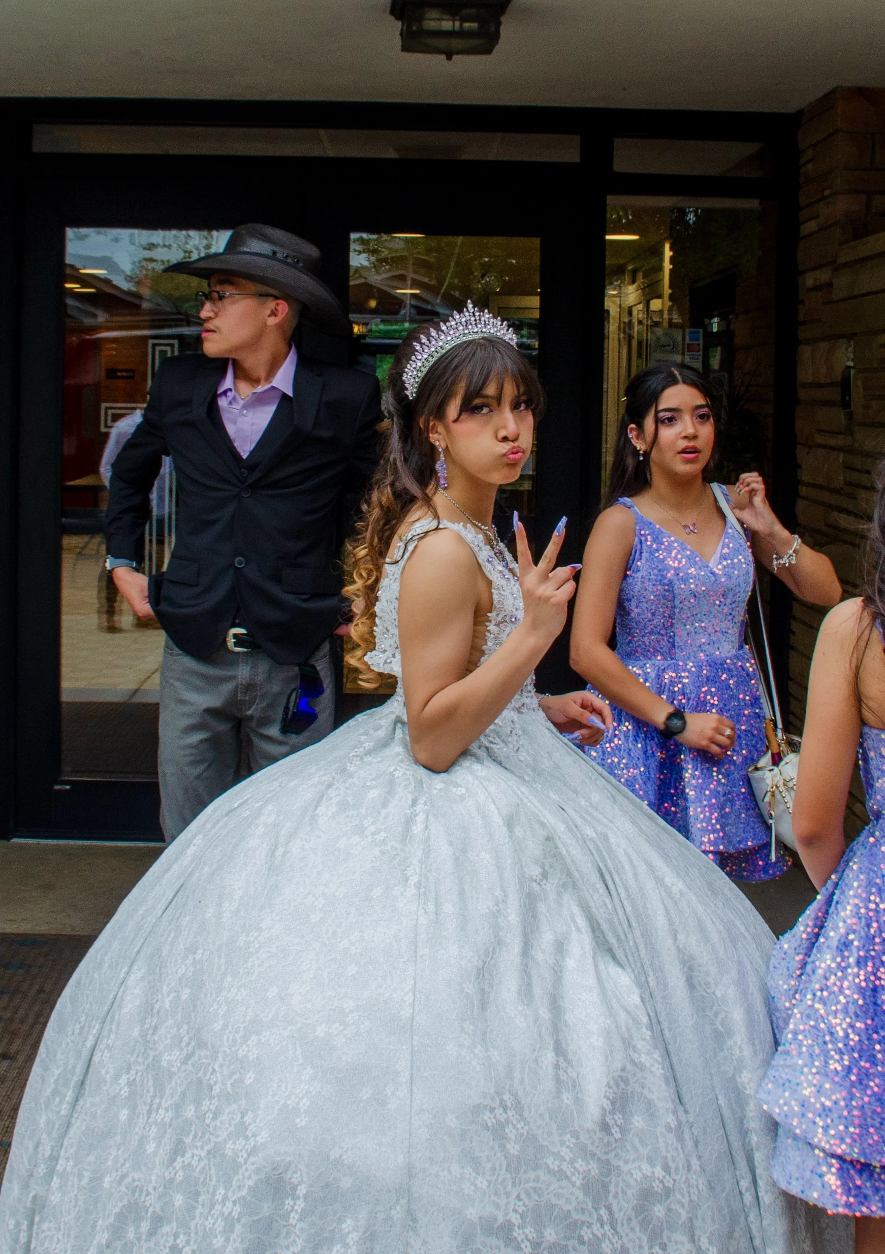 Young woman in a white lace ball gown and tiara making a peace sign, standing next to another young woman in a purple sequin dress, with two other men in dark suits and hats in the background.