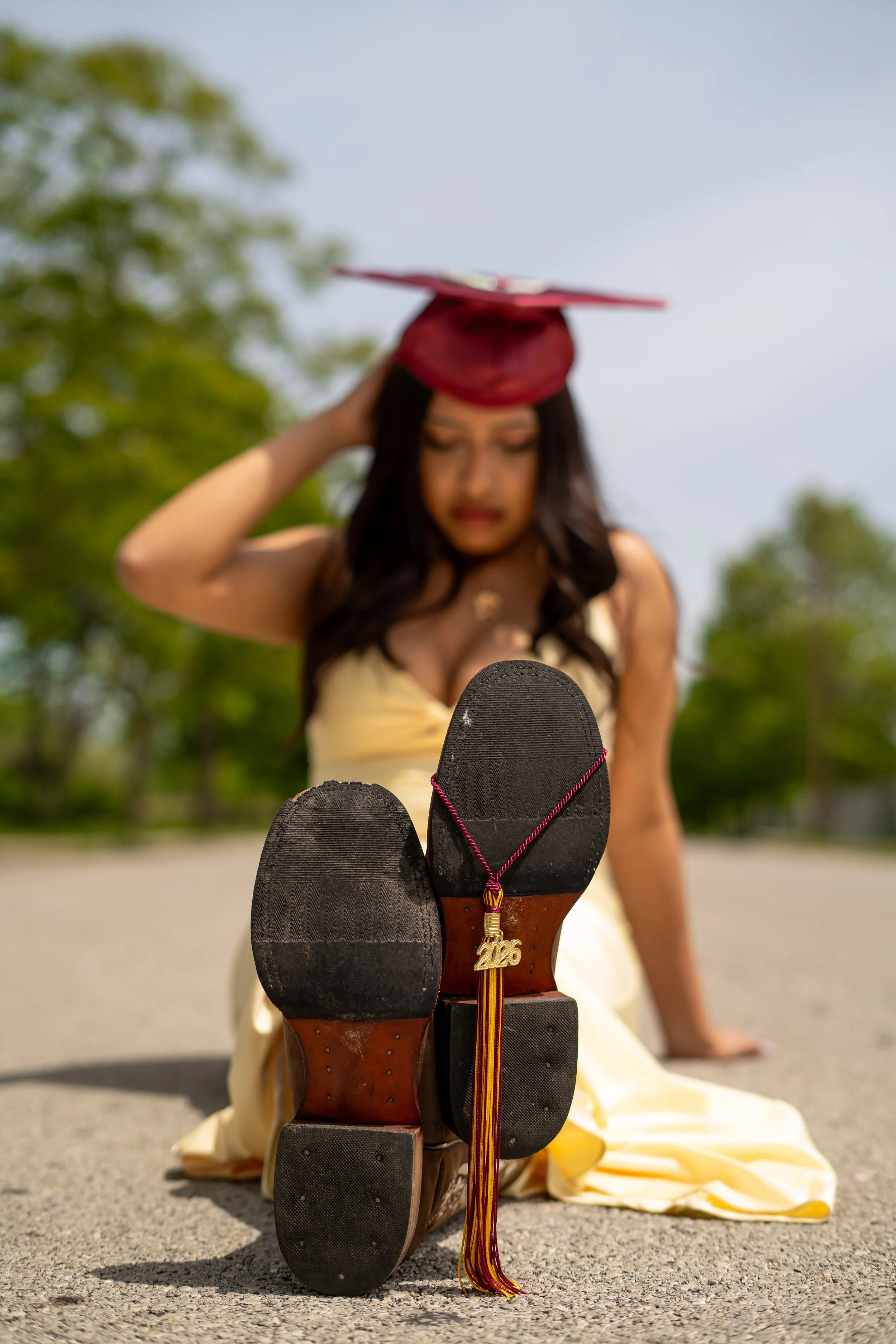 A woman's graduation cap is resting on her head and her shoes are kicked up in the foreground. She is sitting outdoors with green trees and a clear sky in the background.