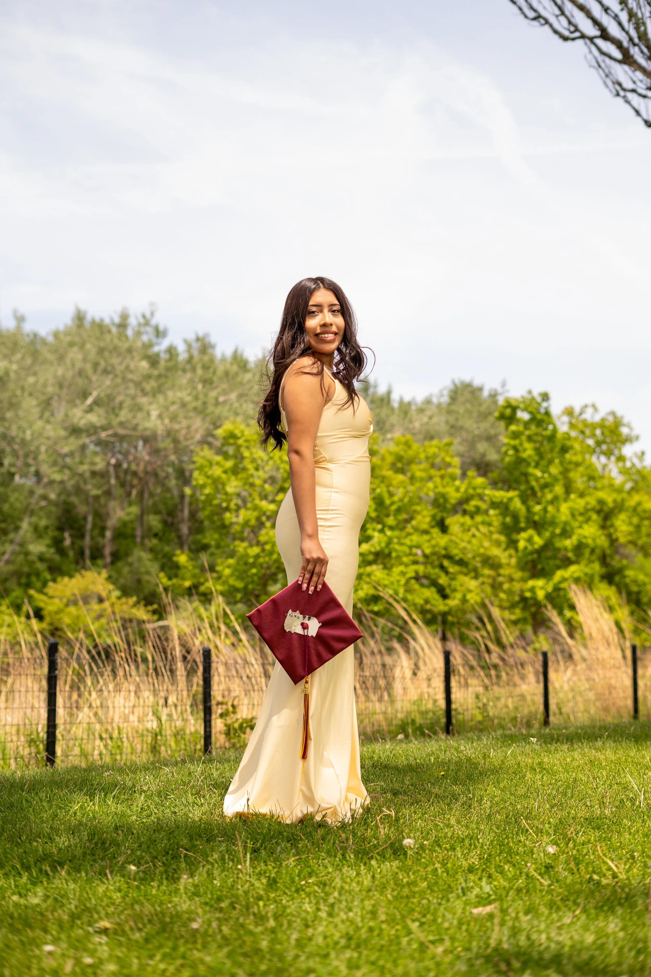 A woman in a cream-colored gown standing on grass in a park, holding a maroon clutch with a decorative pin, smiling, with green trees and a cloudy sky in the background.