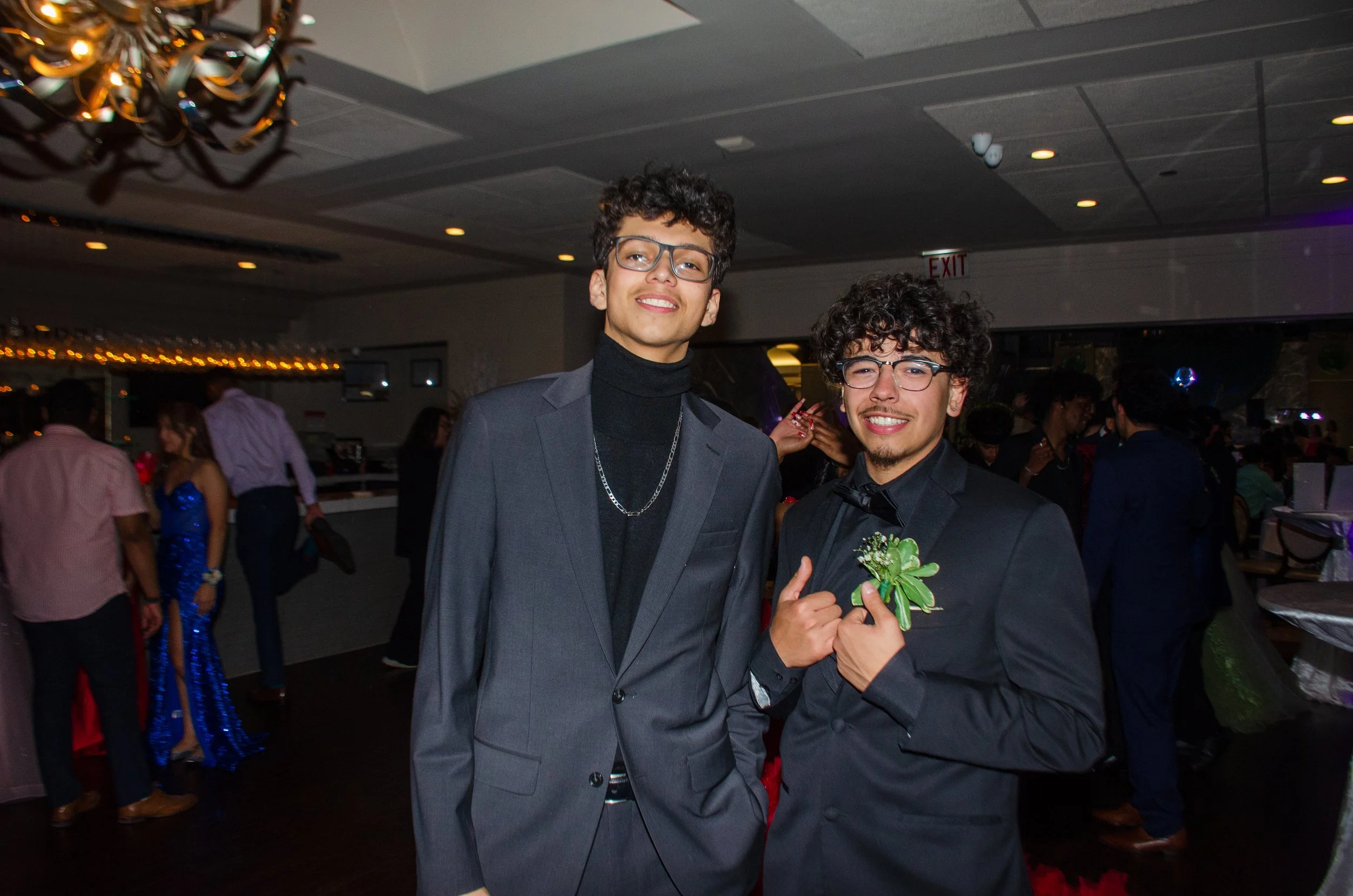 Two young men dressed in formal suits posing at a party with other guests in the background. One is holding a boutonniere.
