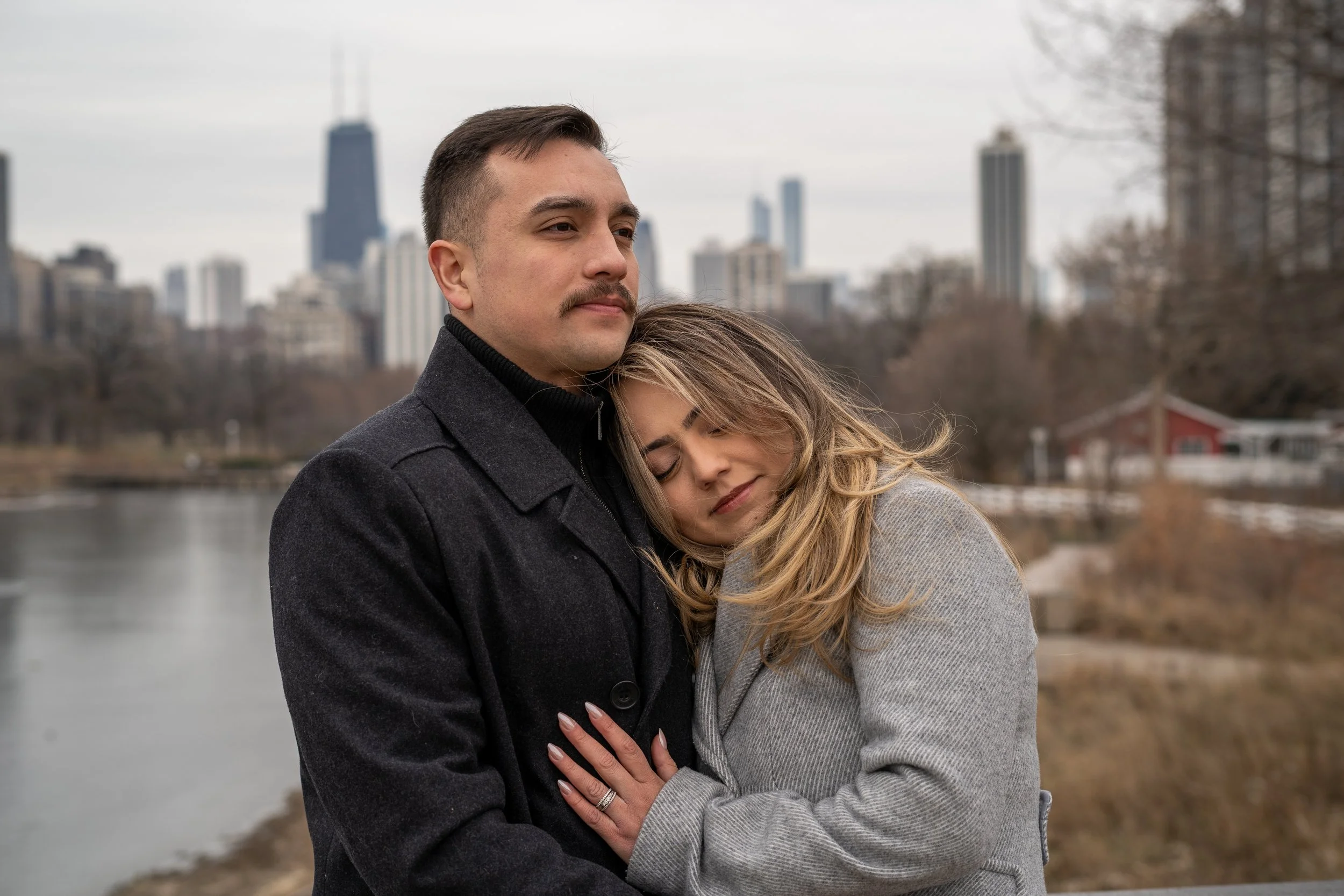 A man and a woman hugging outdoors near a body of water with a city skyline in the background on a cloudy day.