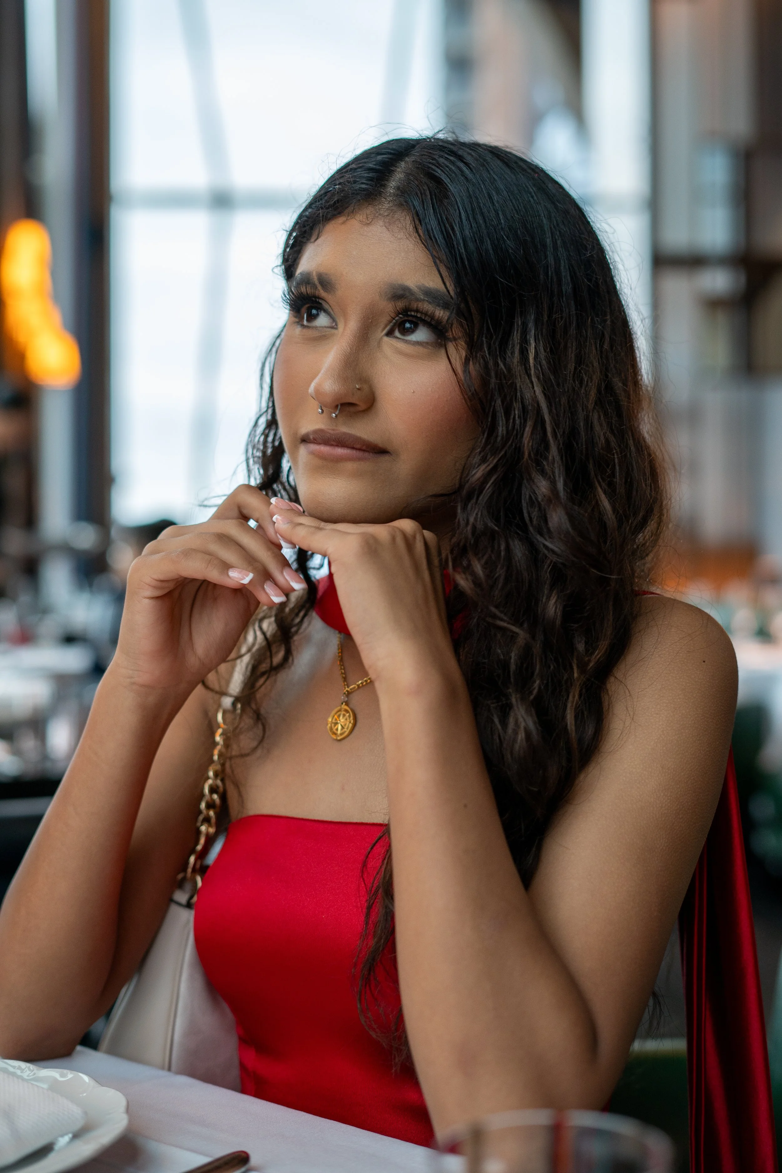 A woman with long dark hair, wearing a red top and gold jewelry, sitting at a table in a restaurant with a window in the background.