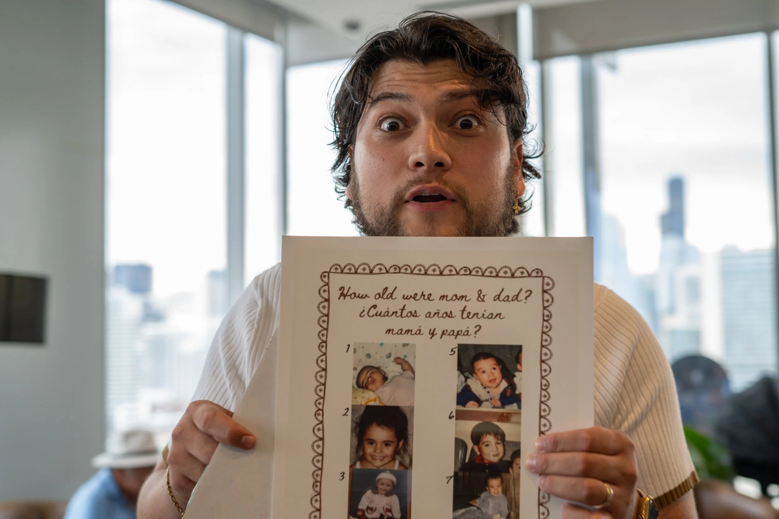 A woman with dark, curly hair and a beard is holding a paper with childhood photos and a question written in English and Spanish. The woman is in an office with large windows and city buildings in the background.