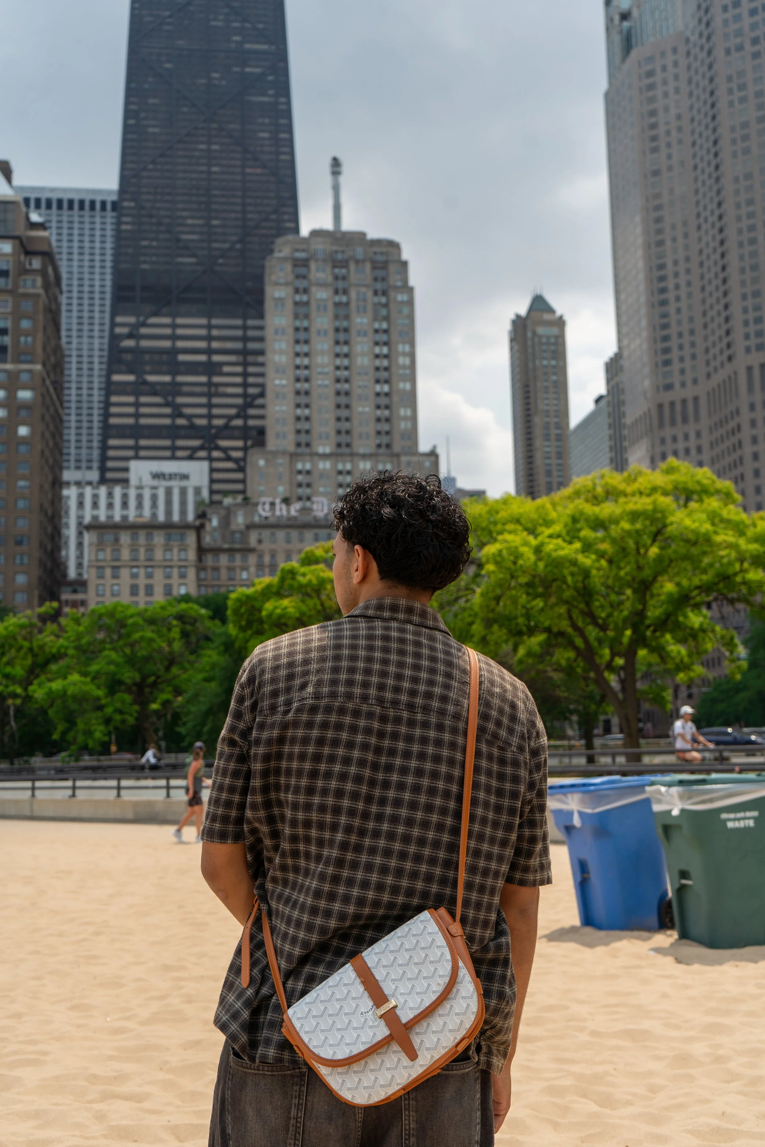 A person with curly hair wearing a checkered shirt and carrying a white and brown bag, standing on a sandy area with trees and skyscrapers in the background.