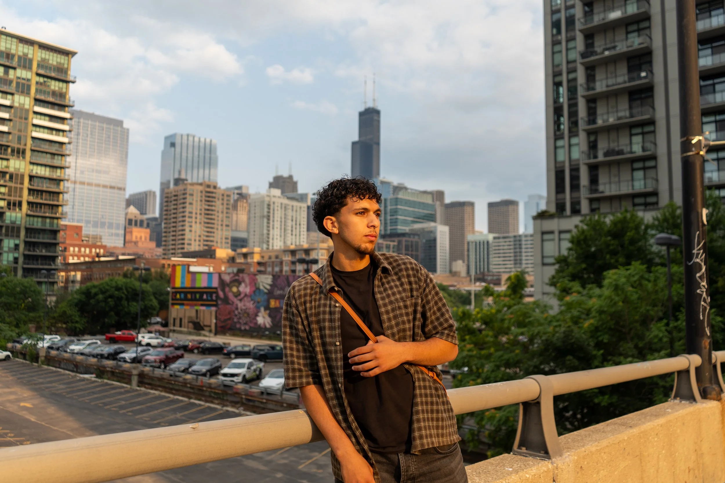 A young man with curly hair and a beard leaning on a bridge railing in an urban cityscape with tall buildings and a parking lot in the background. He is wearing a checkered shirt over a black t-shirt.