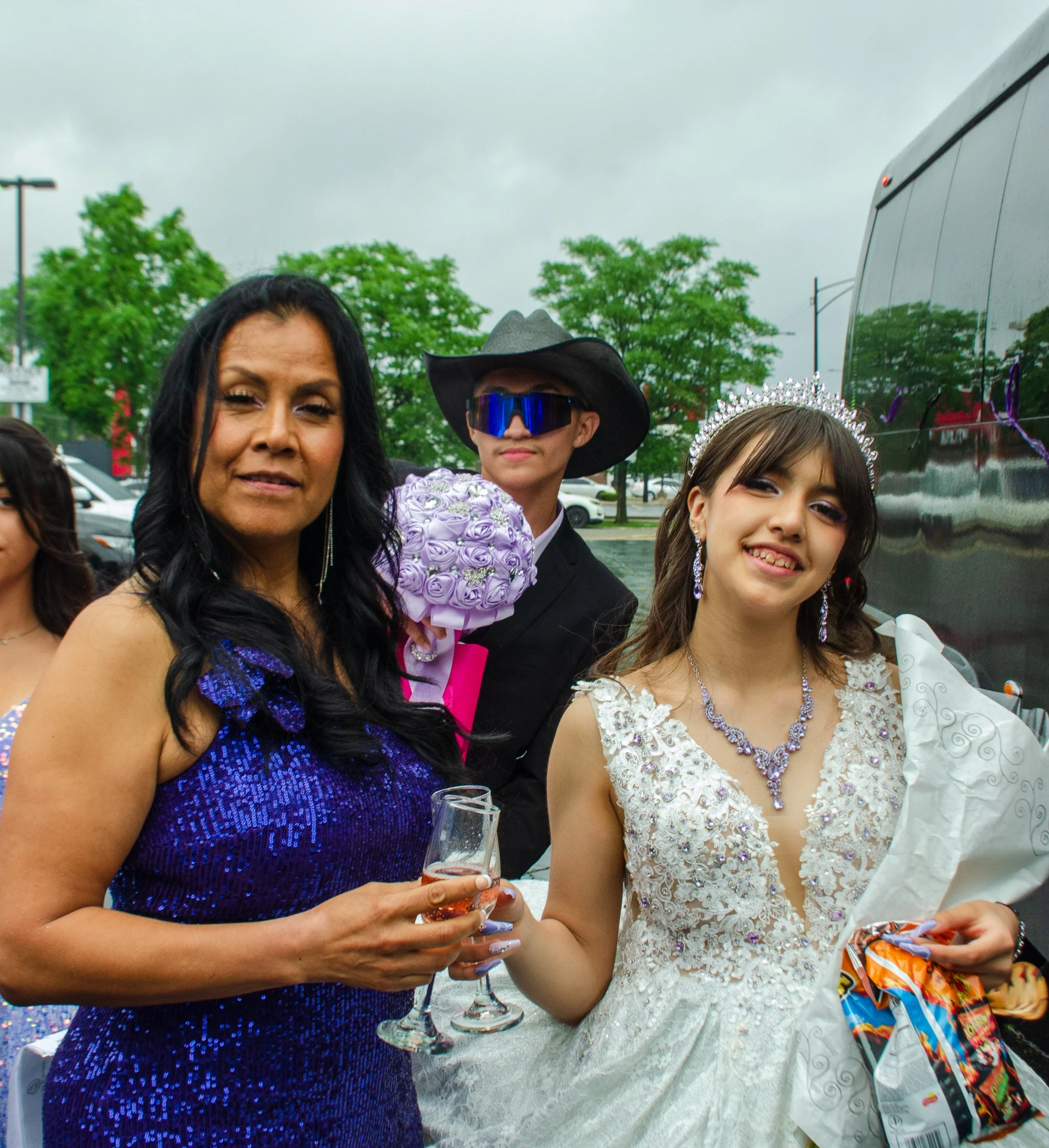 Group of women and girl at a celebration, dressed in formal attire and jewelry, with one holding a glass of wine and others holding snack bags, outdoors on a cloudy day.
