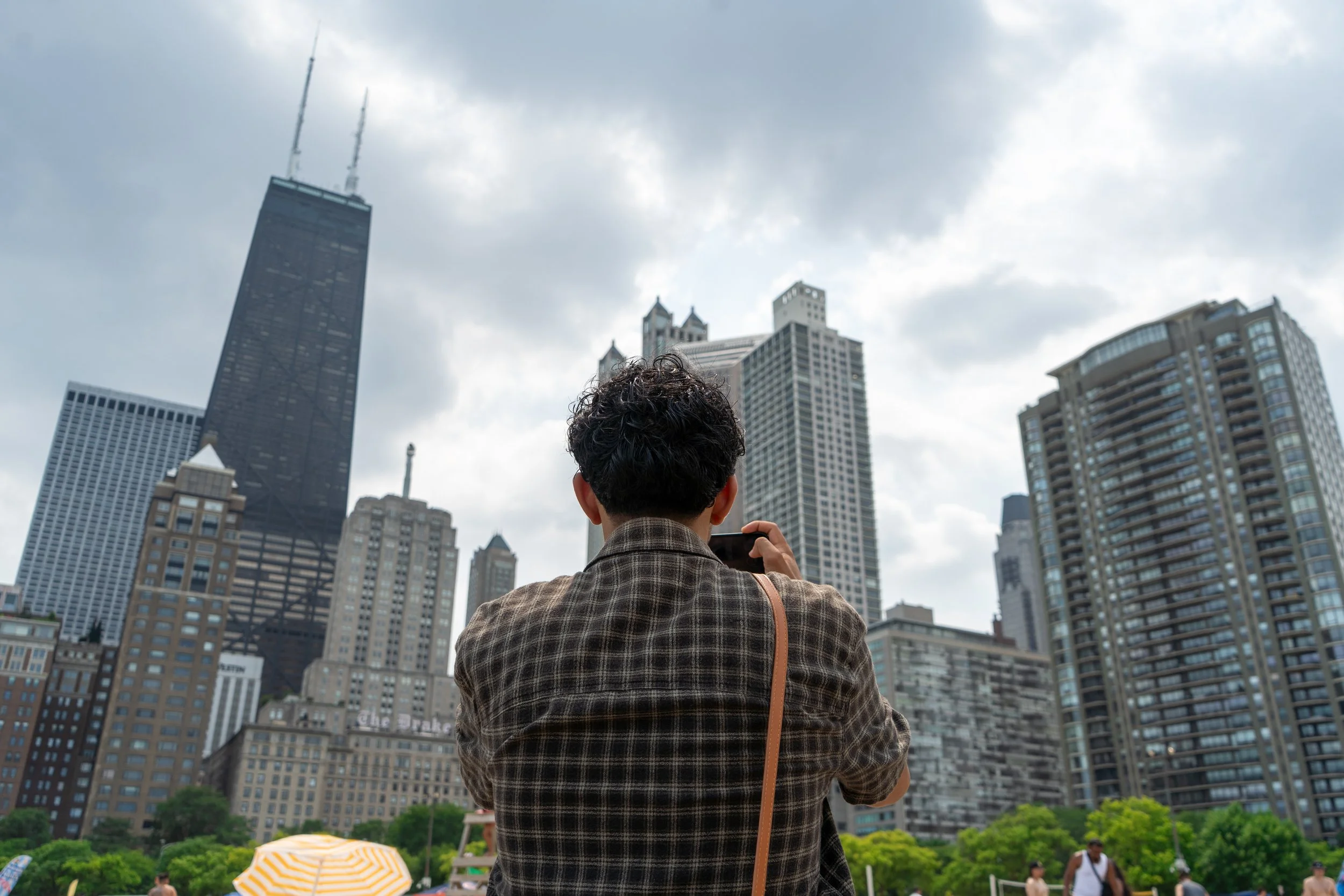 A person with dark, curly hair taking a photo of the Chicago skyline with tall buildings, including the John Hancock Center, during cloudy weather.