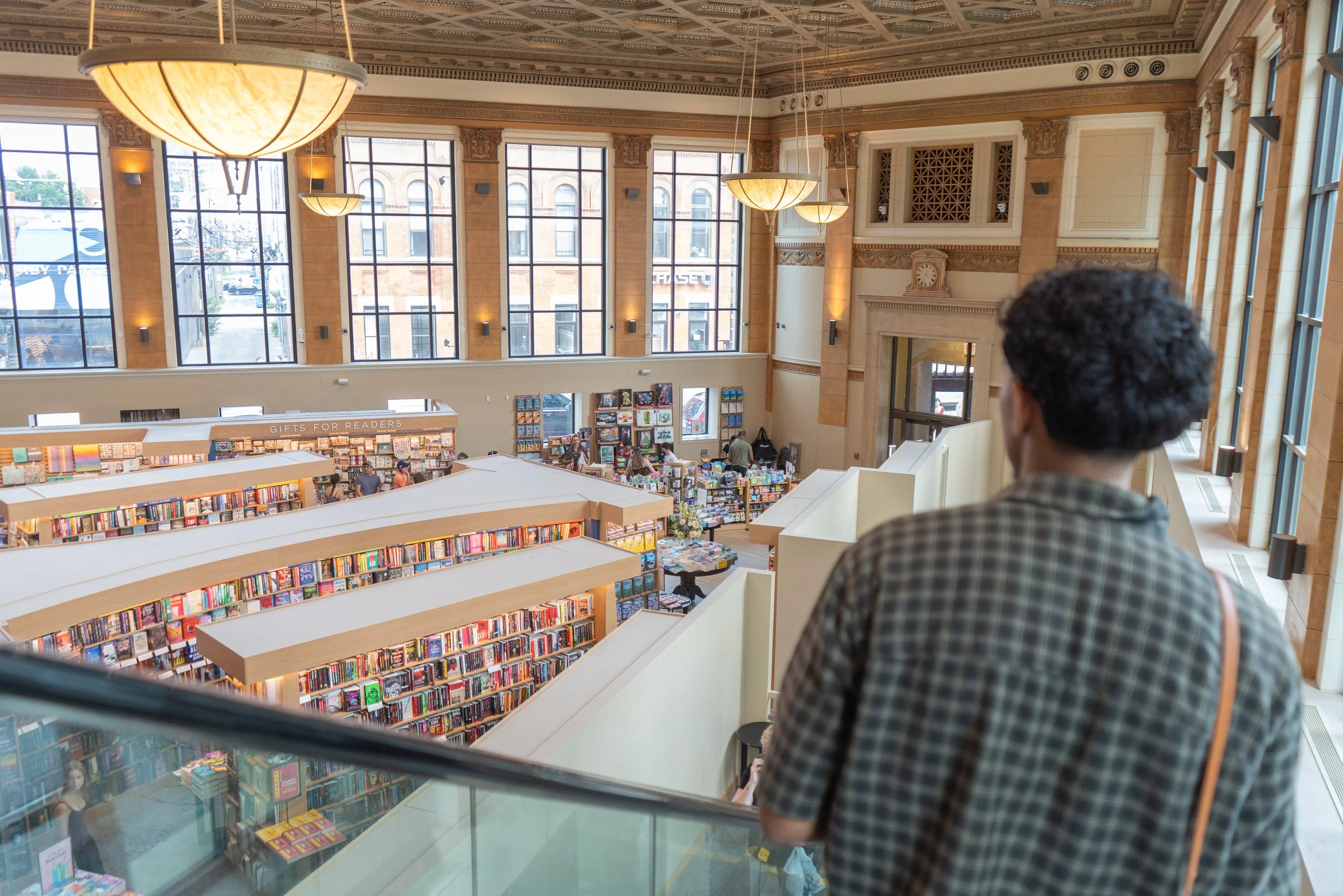 A person with short dark hair wearing a checkered shirt standing inside a large bookstore or library, looking towards the shelves filled with books. The interior features ornate architectural details, large windows, and hanging light fixtures.