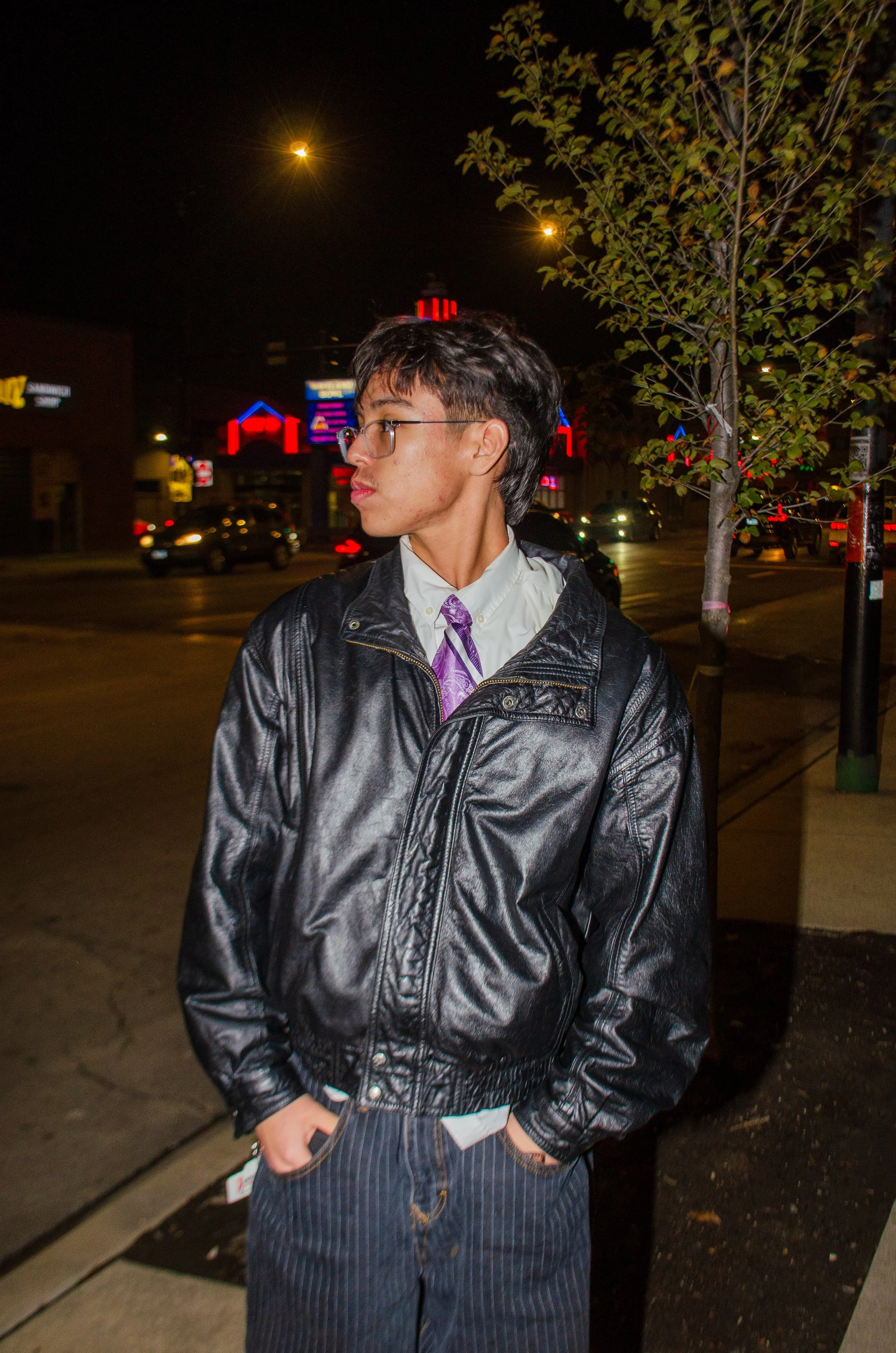 A young man with glasses and short dark hair wearing a white shirt, purple tie, black leather jacket, and pinstriped pants standing on a city sidewalk at night with cars and brightly lit signs in the background.