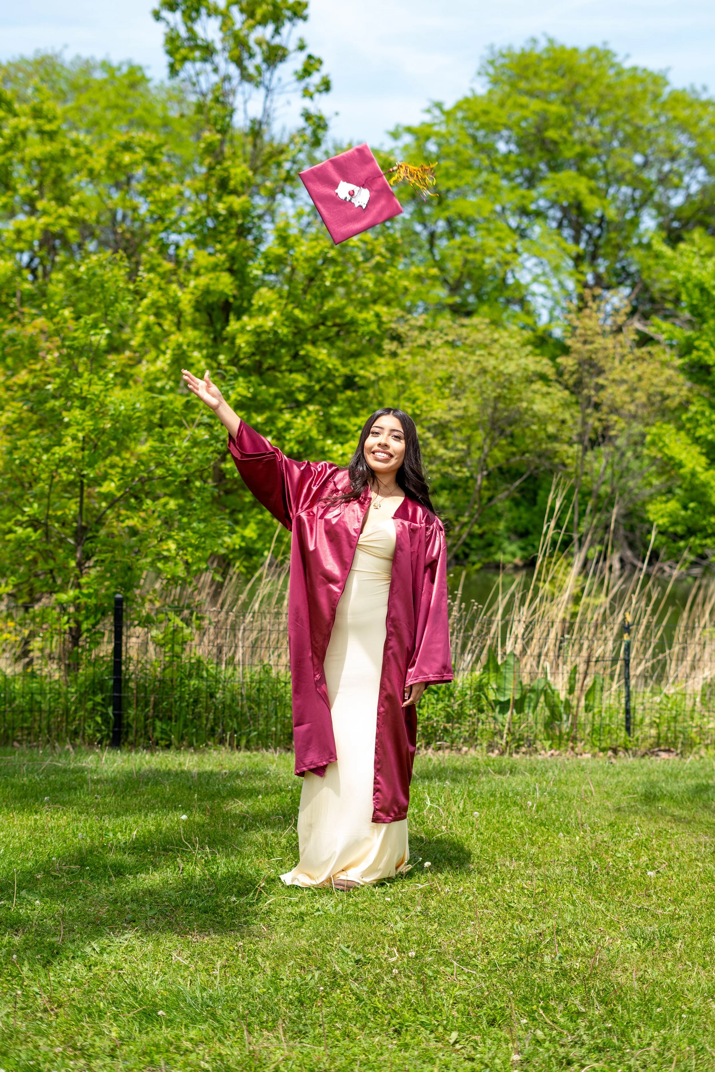 A woman in a white dress and maroon graduation gown celebrating outdoors, throwing her cap in the air with trees and blue sky in the background.