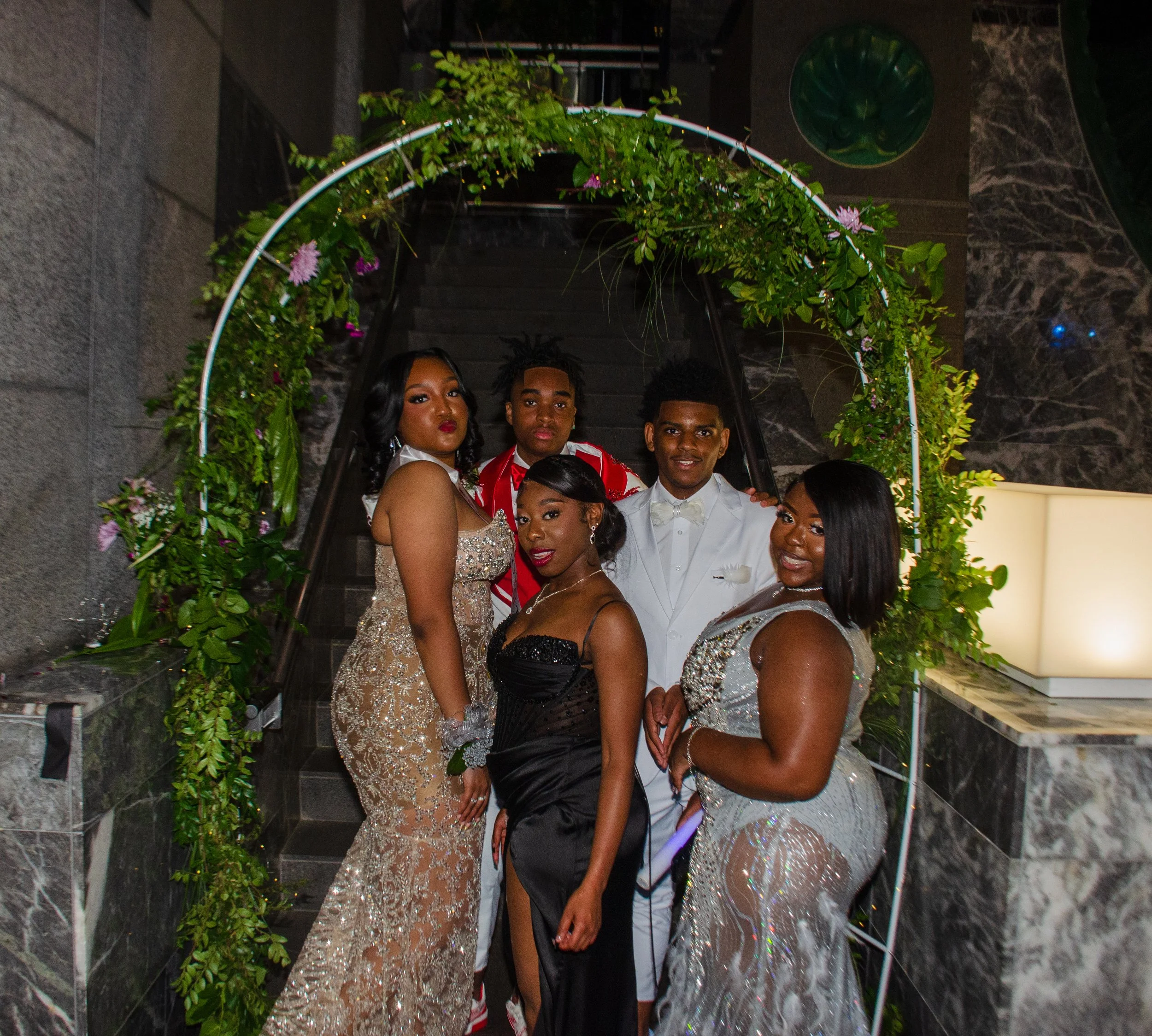 A group of young people dressed in formal attire under a decorated floral arch, posing for a photo at an indoor event.