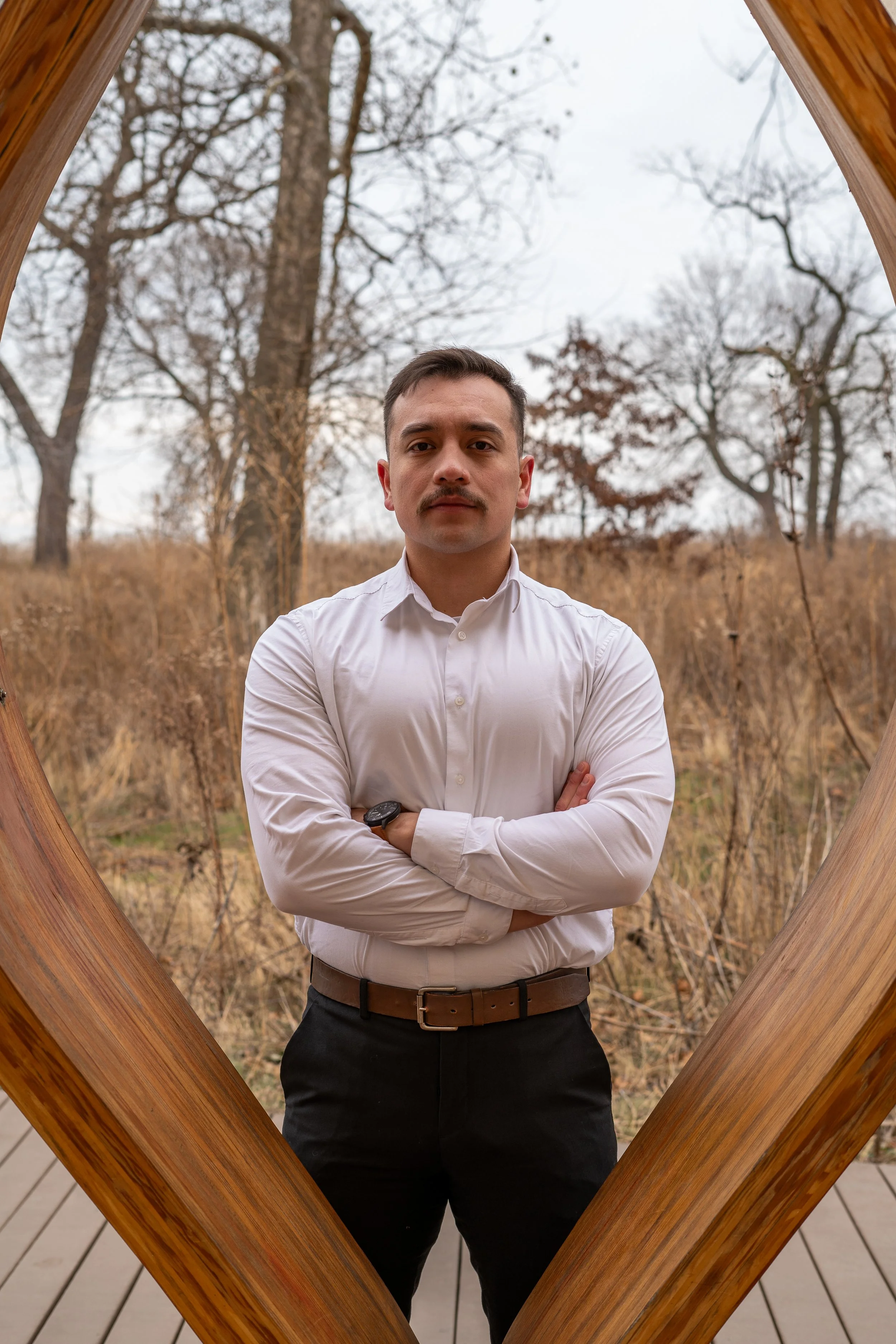 A man wearing a white dress shirt and black pants standing outdoors during fall with bare trees behind him, framed by a large wooden diamond-shaped structure.