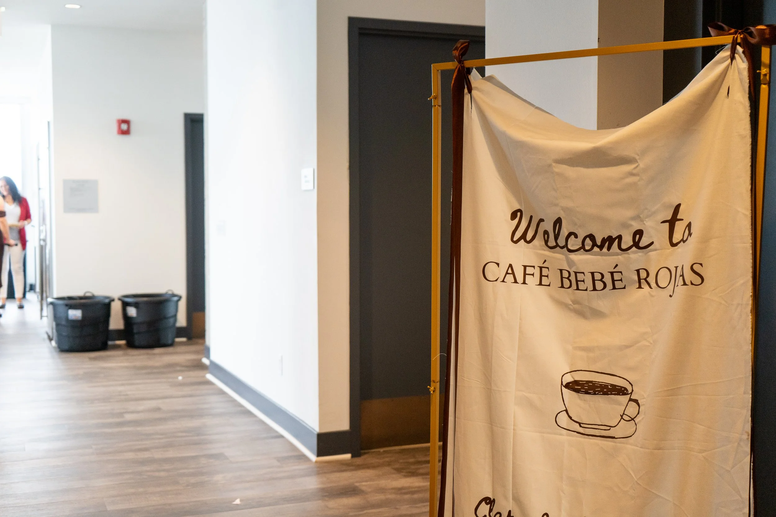 A sign welcoming visitors to Café Bebé Rojas, with a simple illustration of a coffee cup, on a fabric banner set up inside a modern building.