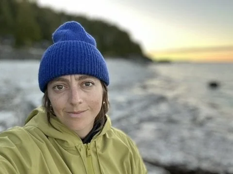 Close-up of a woman in a blue beanie and yellow jacket taking a selfie near a body of water during sunset.