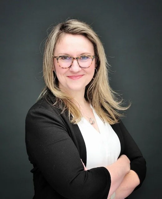 A woman with shoulder-length blonde hair and glasses, wearing a black blazer over a white top, smiling with arms crossed in front of a dark background.