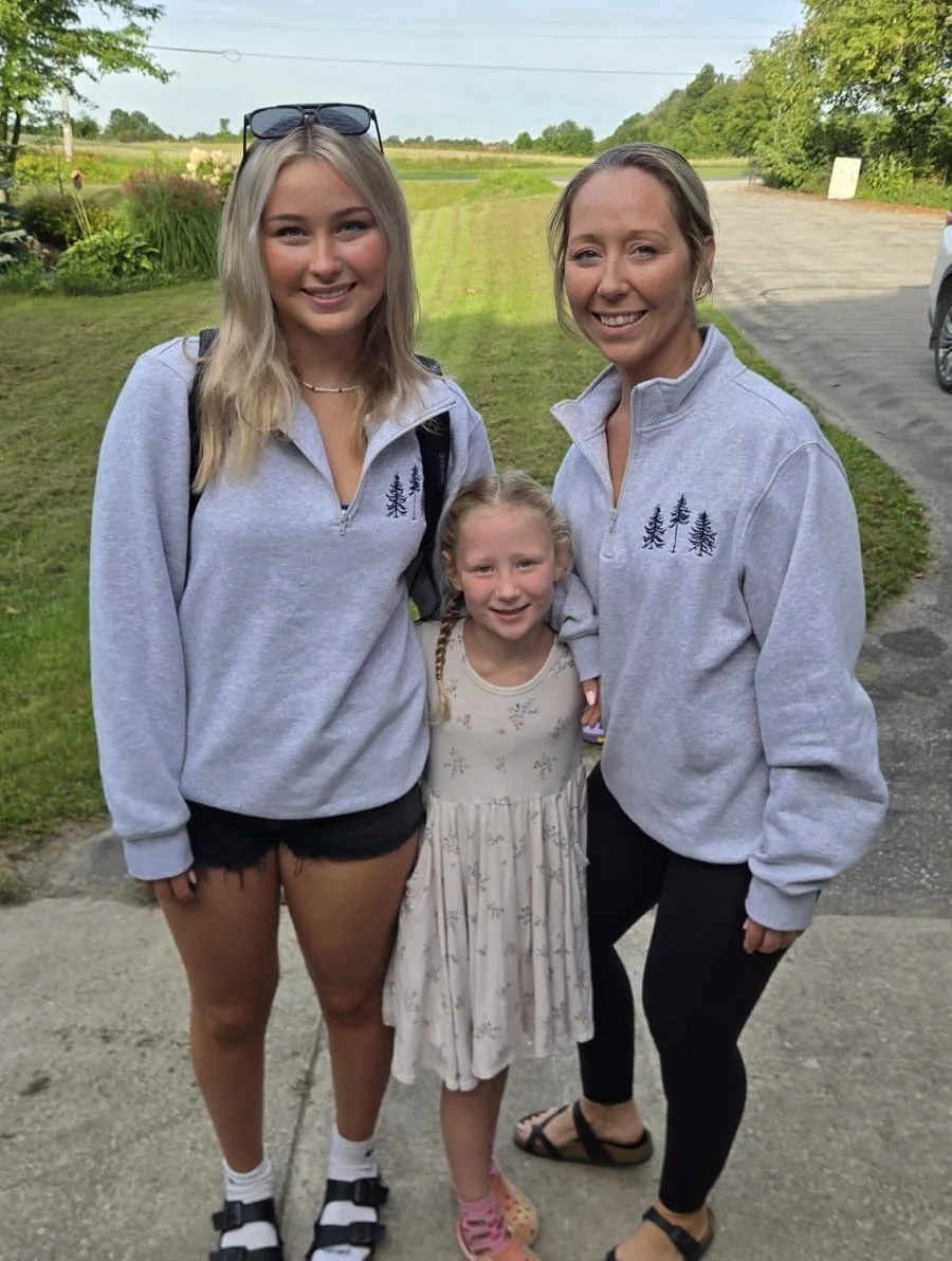 Three females smiling outdoors on a grassy area with trees in the background. Two older females and a young girl, all dressed in casual clothing. The girls are wearing light gray sweatshirts with tree designs.