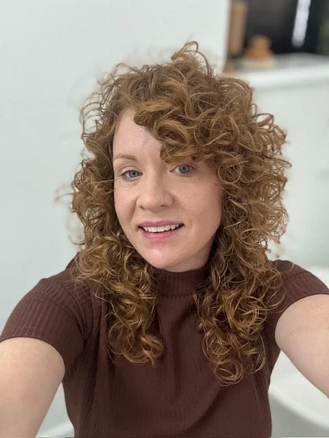 A woman with curly red hair taking a selfie in a room with light-colored walls and blurry furniture in the background.