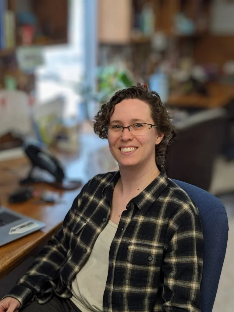 A young woman with short curly hair, glasses, and a checkered shirt sitting at a desk in an office environment, smiling at the camera.