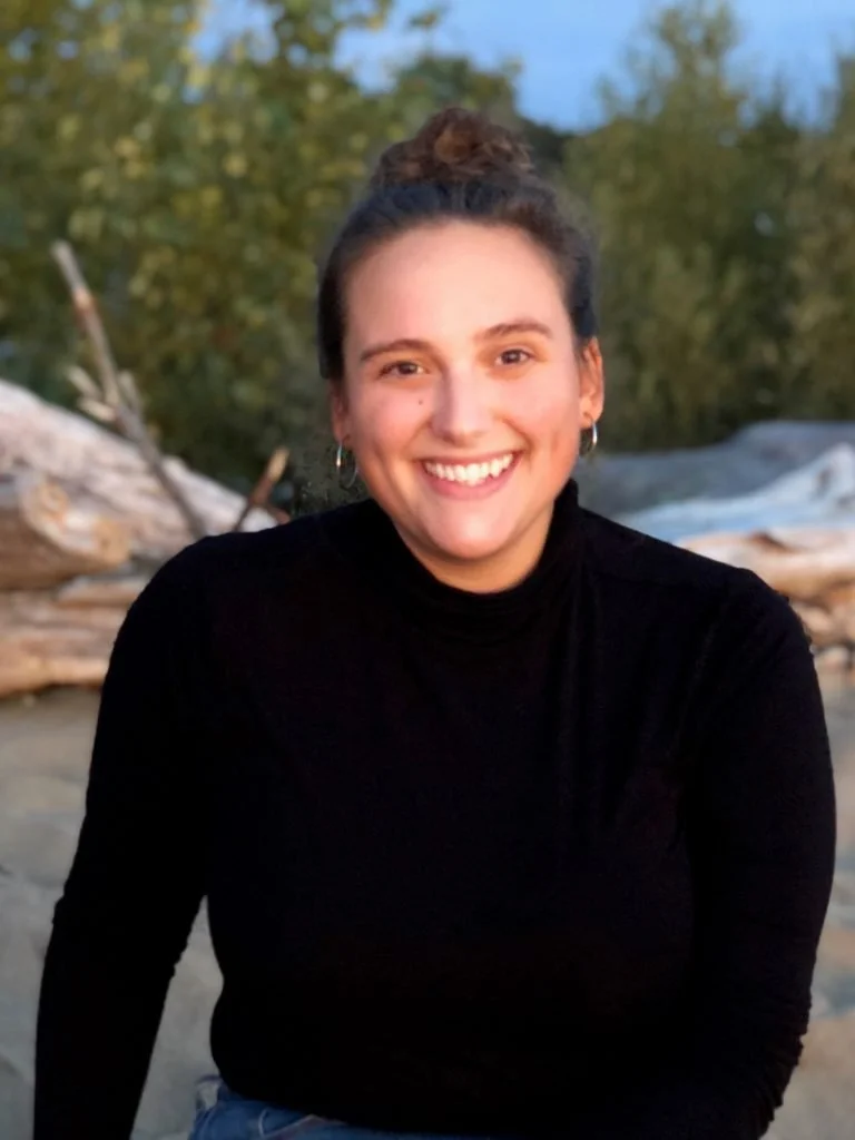 A young woman with light skin, dark hair tied up, wearing hoop earrings and a black turtleneck, smiling outdoors near a beach with driftwood and trees in the background.