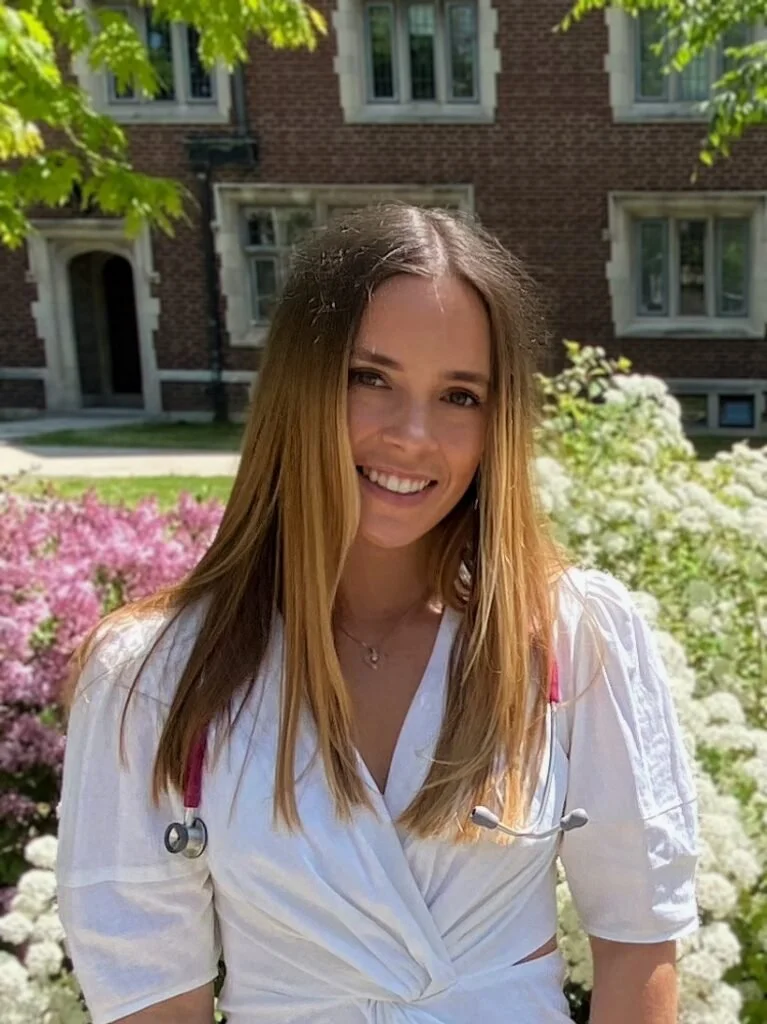 A young woman with long brown hair smiling outdoors, wearing a white medical uniform with a pink stethoscope, standing in front of a garden with pink and white flowers and a brick building in the background.