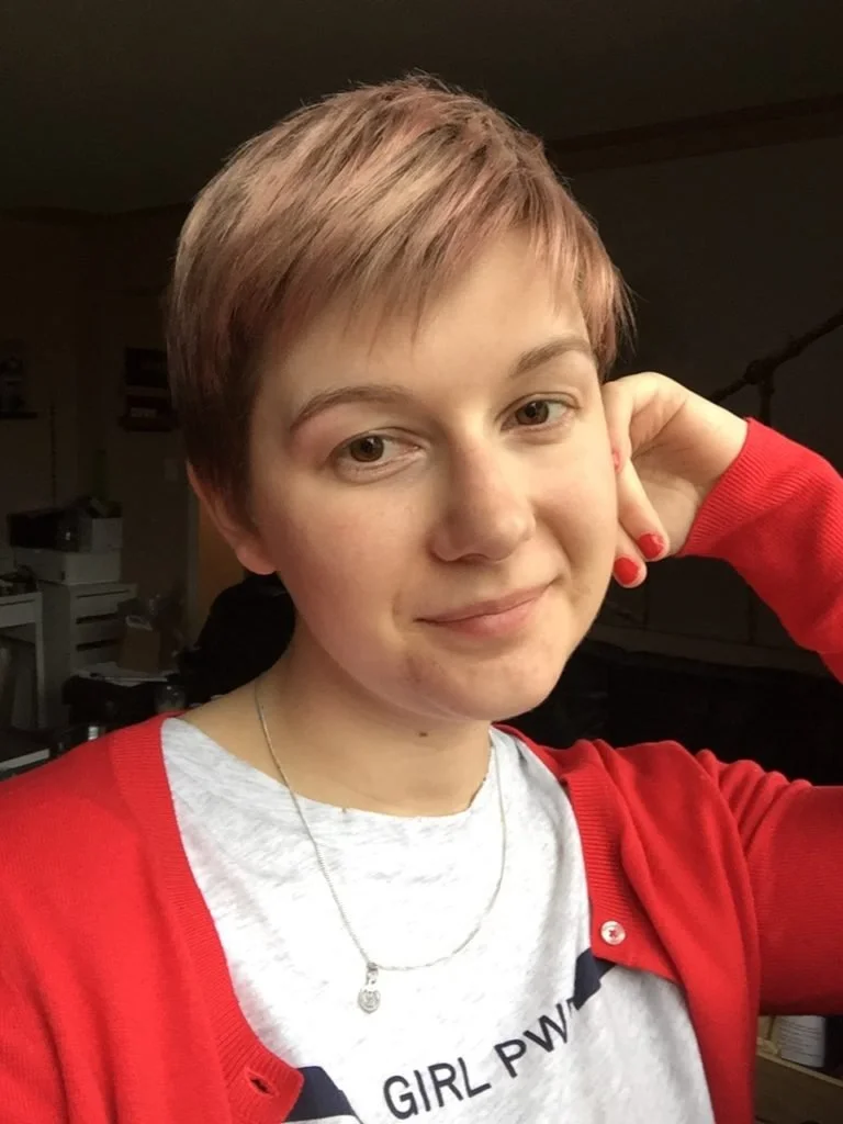 Portrait of a young woman with short, light brown hair, wearing a red cardigan and a white t-shirt, smiling softly and posing with one hand behind her head in an indoor setting.