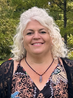 A woman with curly, shoulder-length gray hair smiling outdoors in a park with trees in the background.