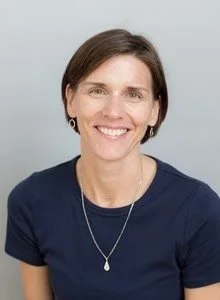 A woman with short dark hair, wearing a navy blue shirt, smiling, against a plain light background.