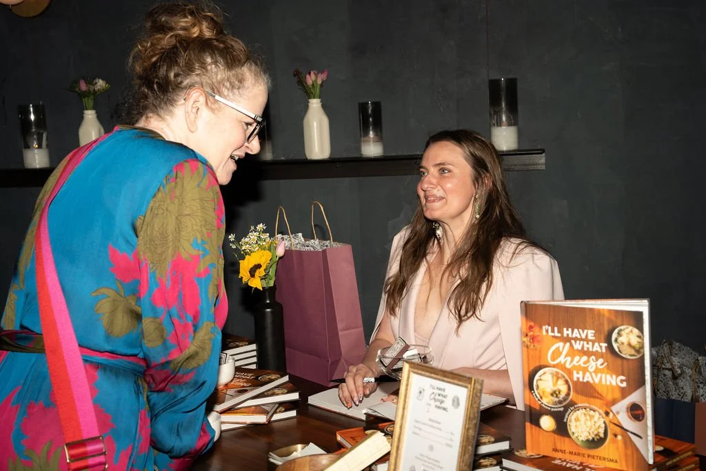 A woman with long brown hair in a light pink blazer is smiling at another woman with curly hair wearing glasses and a colorful floral jacket. They are interacting above a table with various books and a framed photo, in a dimly lit room with a dark wall and vases with flowers in the background.