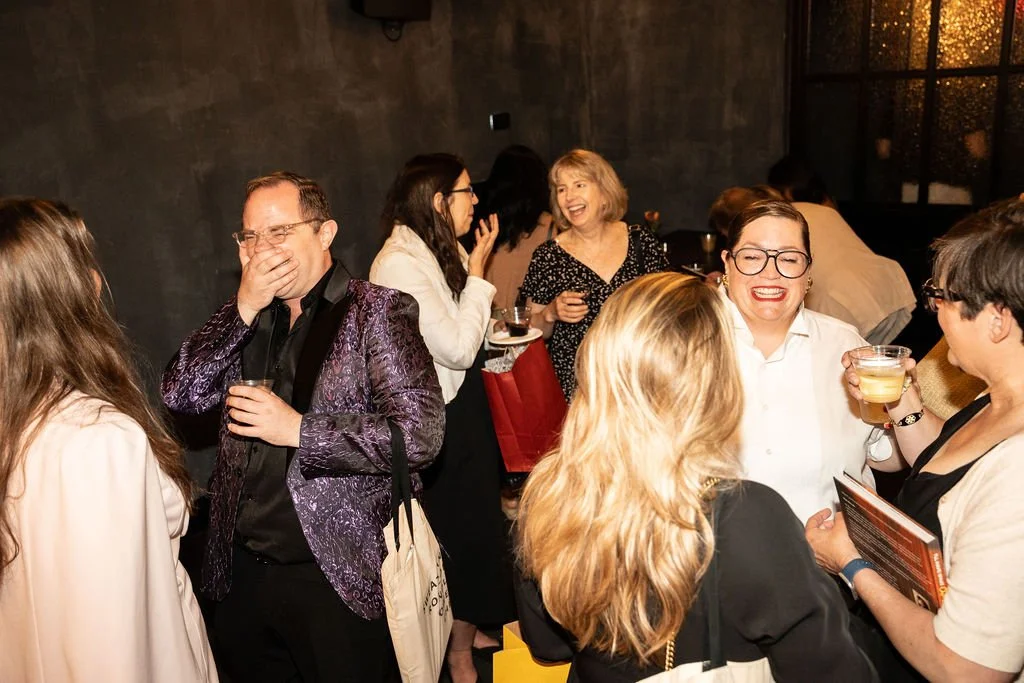 Group of people laughing and talking at an indoor social gathering or party, holding drinks, in a dimly lit room.