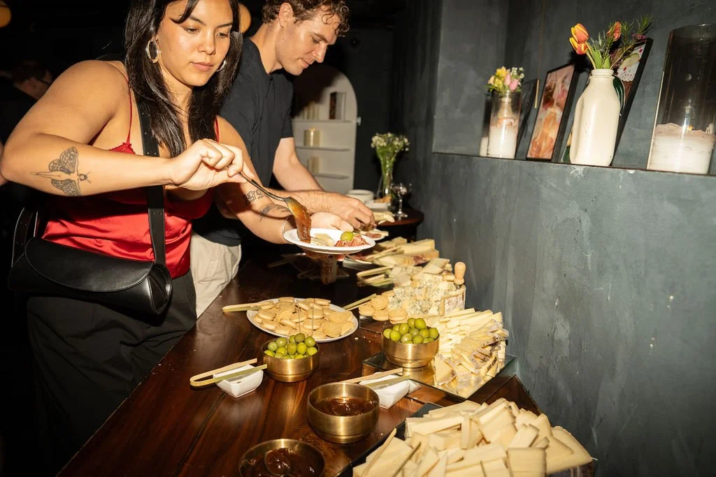 Two people at a cheese and fruit buffet, with slices of cheese, bowls of green olives, and other appetizers on a wooden table.