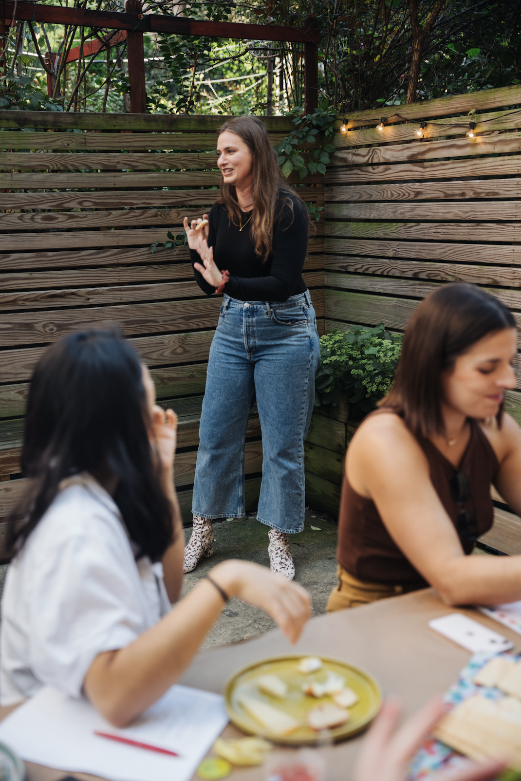 A woman standing outdoors near a wooden fence, speaking to a group of seated women, with greenery and string lights in the background.