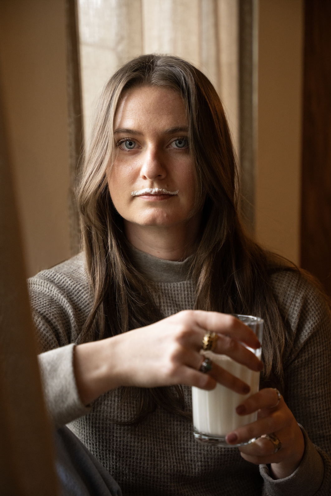 A woman with long brown hair and blue eyes holding a glass of milk, wearing a beige sweater, sitting indoors with curtains in the background.