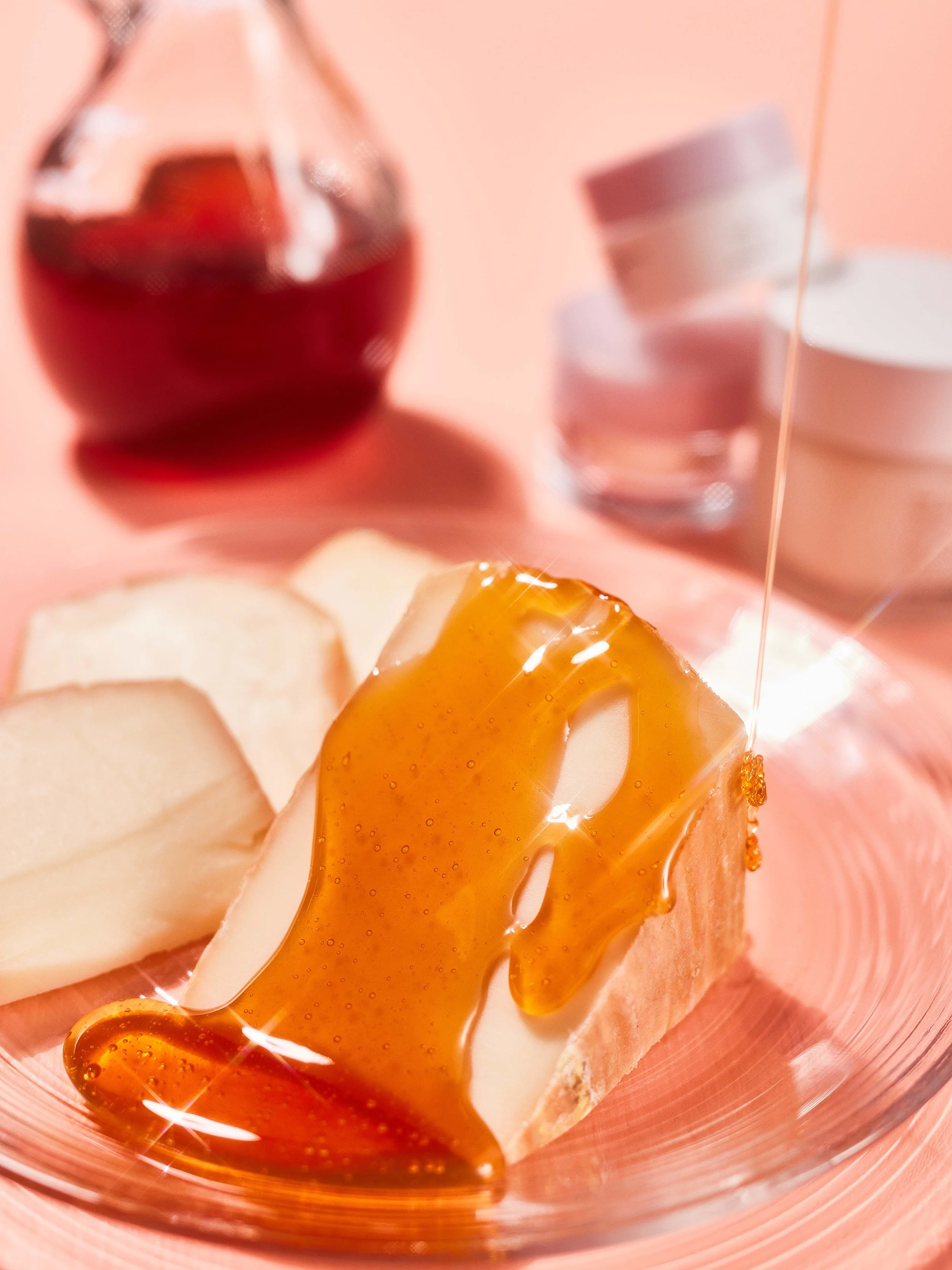 Close-up of a slice of bread with honey drizzling over it, with a glass of red wine and some stacked round objects in the background.