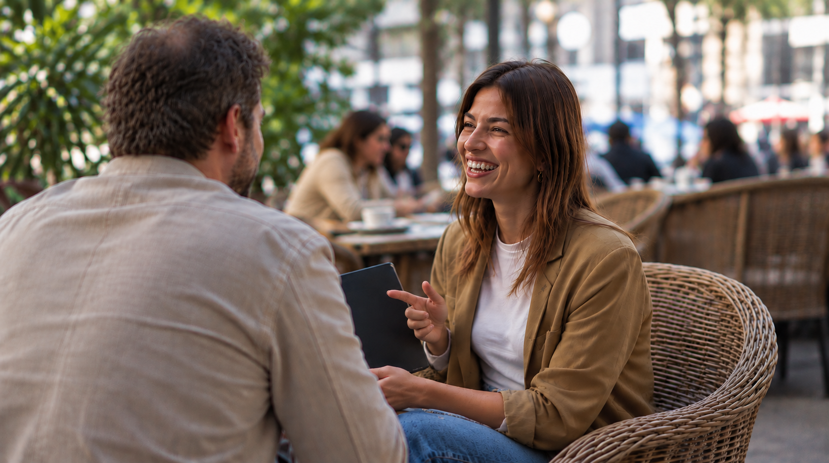 A woman with brown hair and a tan jacket smiling and pointing to a man with brown hair and a beige shirt while sitting at an outdoor cafe.