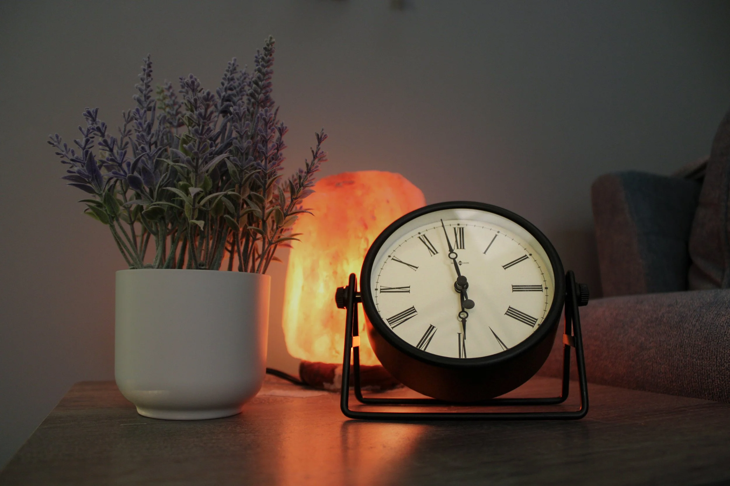 A table with a potted plant, a salt lamp, and a black clock showing 11:03, set against a plain wall.