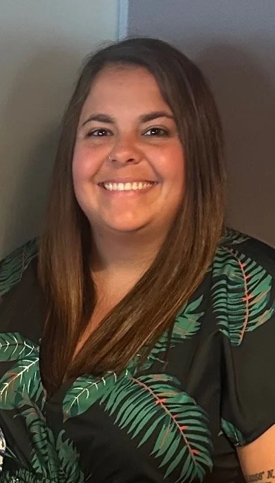 A woman with straight brown hair smiling at the camera, wearing a black shirt with green leaf patterns.