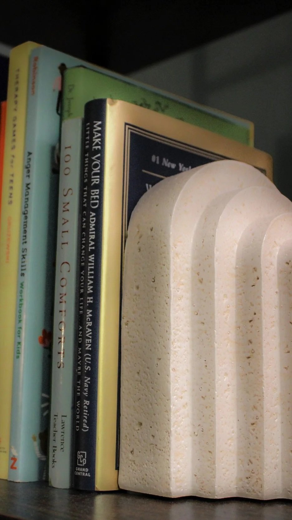 Books on a shelf with a decorative white sculpture in front. The books include titles about management skills, US Navy history, and others. The sculpture has a textured, porous surface with arch shapes.